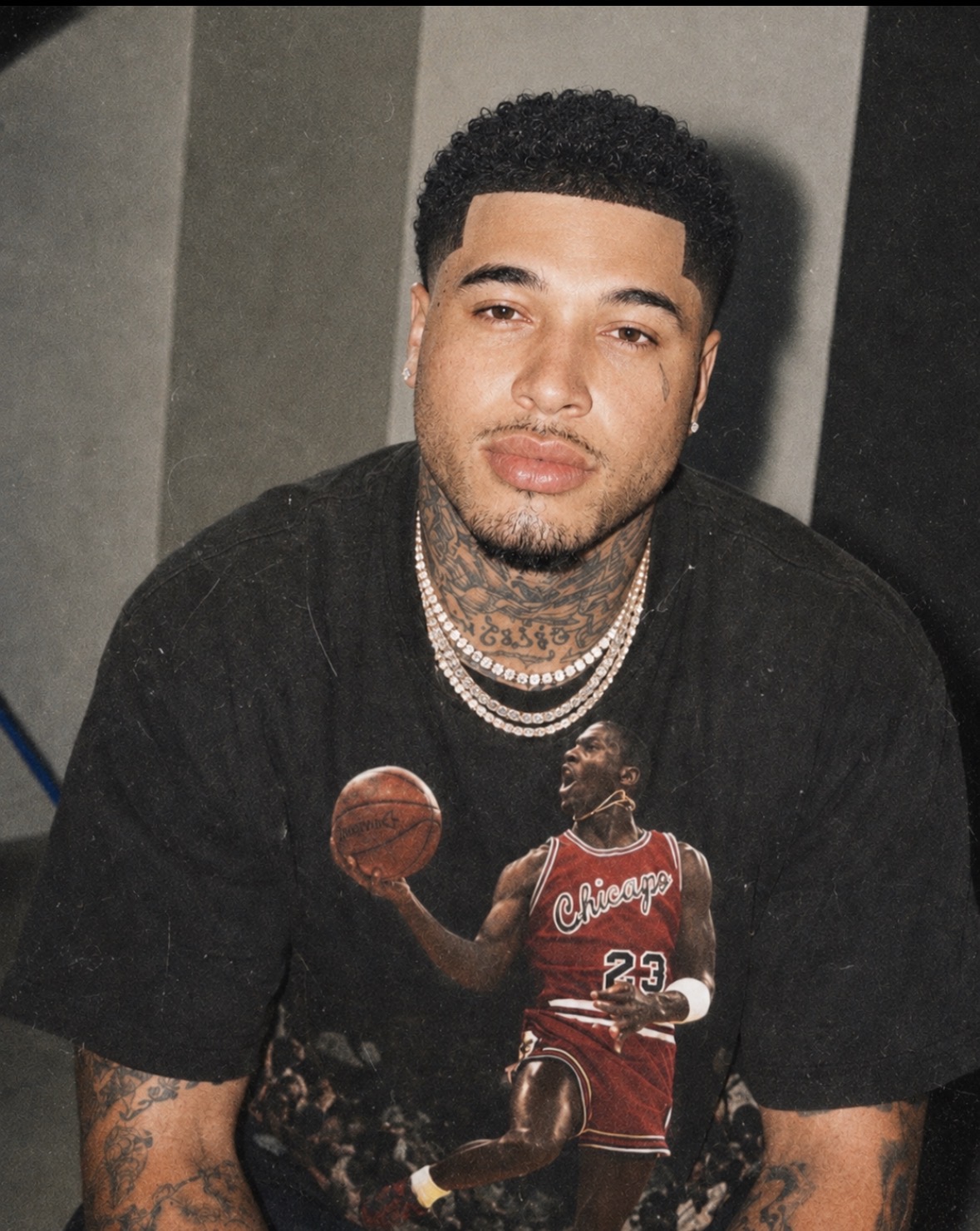 Young man with tattoos and jewelry, wearing a black shirt, sitting indoors with a neutral background.