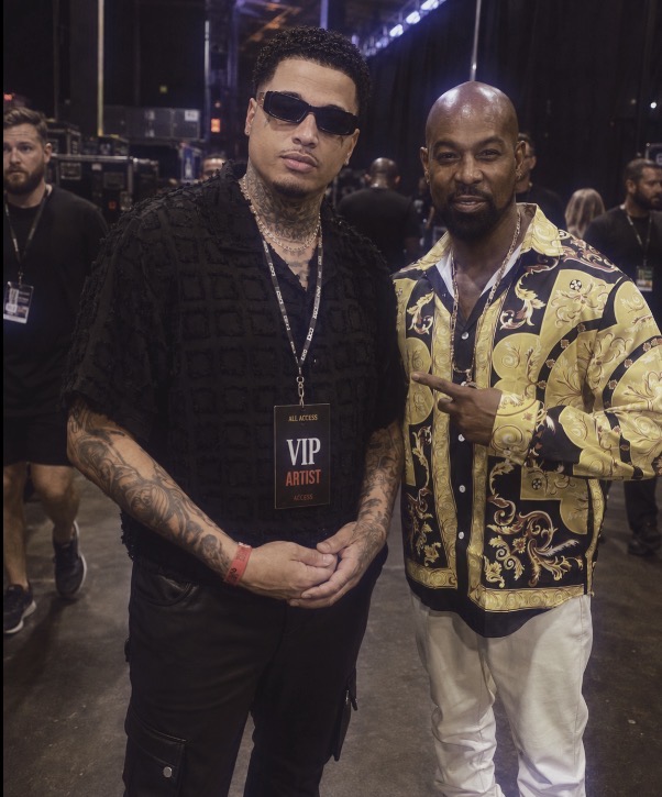 Two men standing together at an event, one wearing sunglasses and a black outfit, the other in a patterned jacket, both smiling.
