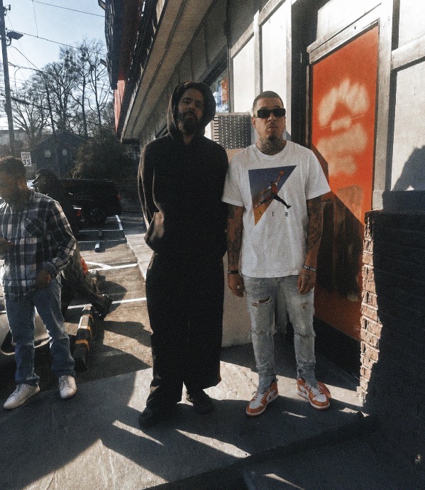 Two men standing outside a building, one wearing a black outfit and the other in a white t-shirt with a graphic, sunglasses, and jeans. Sunlight casts shadows.