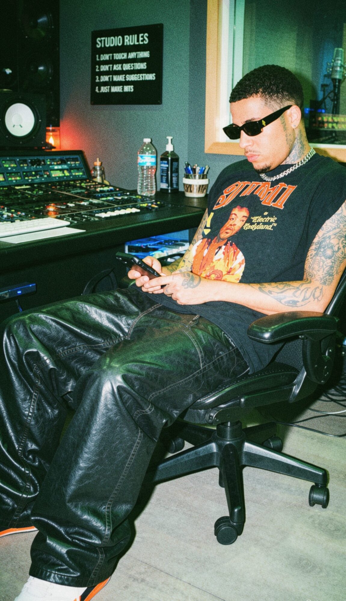Person with sunglasses and tattoos sitting in a chair, using a phone, in a room with music equipment and bottles on a table.