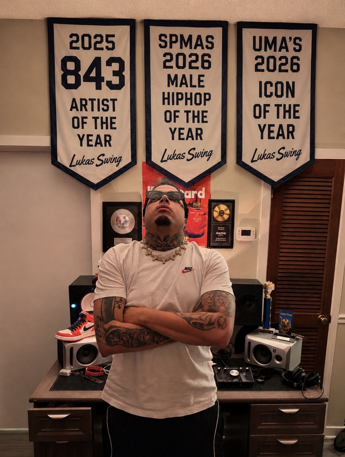 Man with tattoos and glasses stands with arms crossed in a room with music equipment and framed awards on the wall.