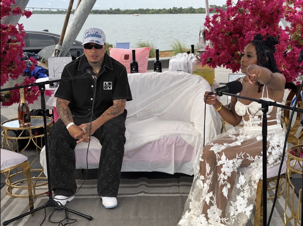 Two people sit on a white couch with pink flowers and a lake in the background, one speaking into a microphone.