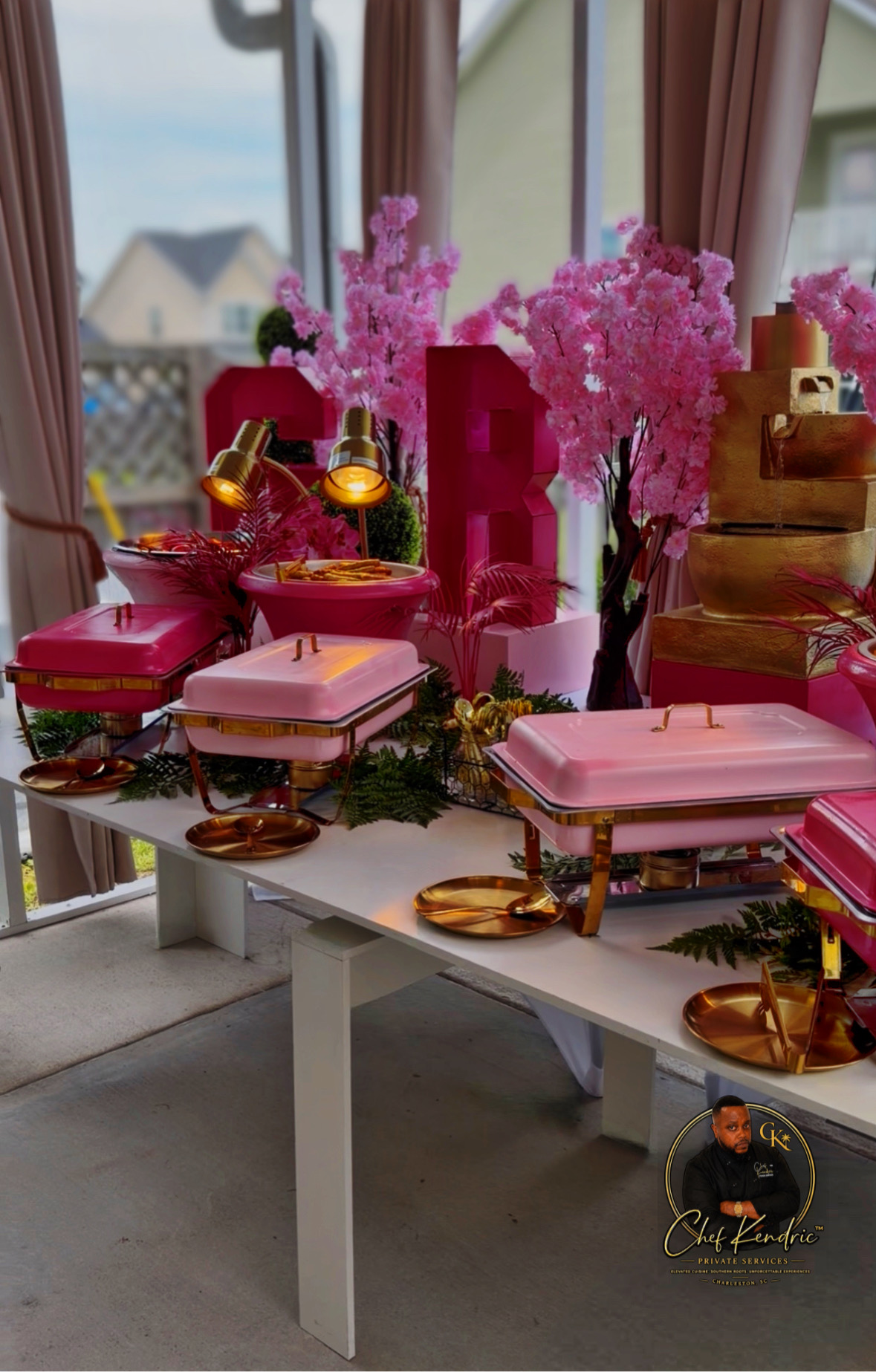 Table decorated with pink and gold accessories, pink flowers, and gift boxes, set near a window with curtains.