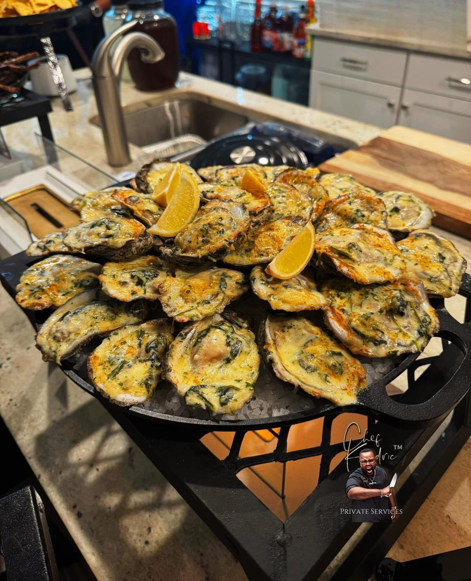 A large pile of cooked oysters garnished with lemon wedges on a black tray in a kitchen.