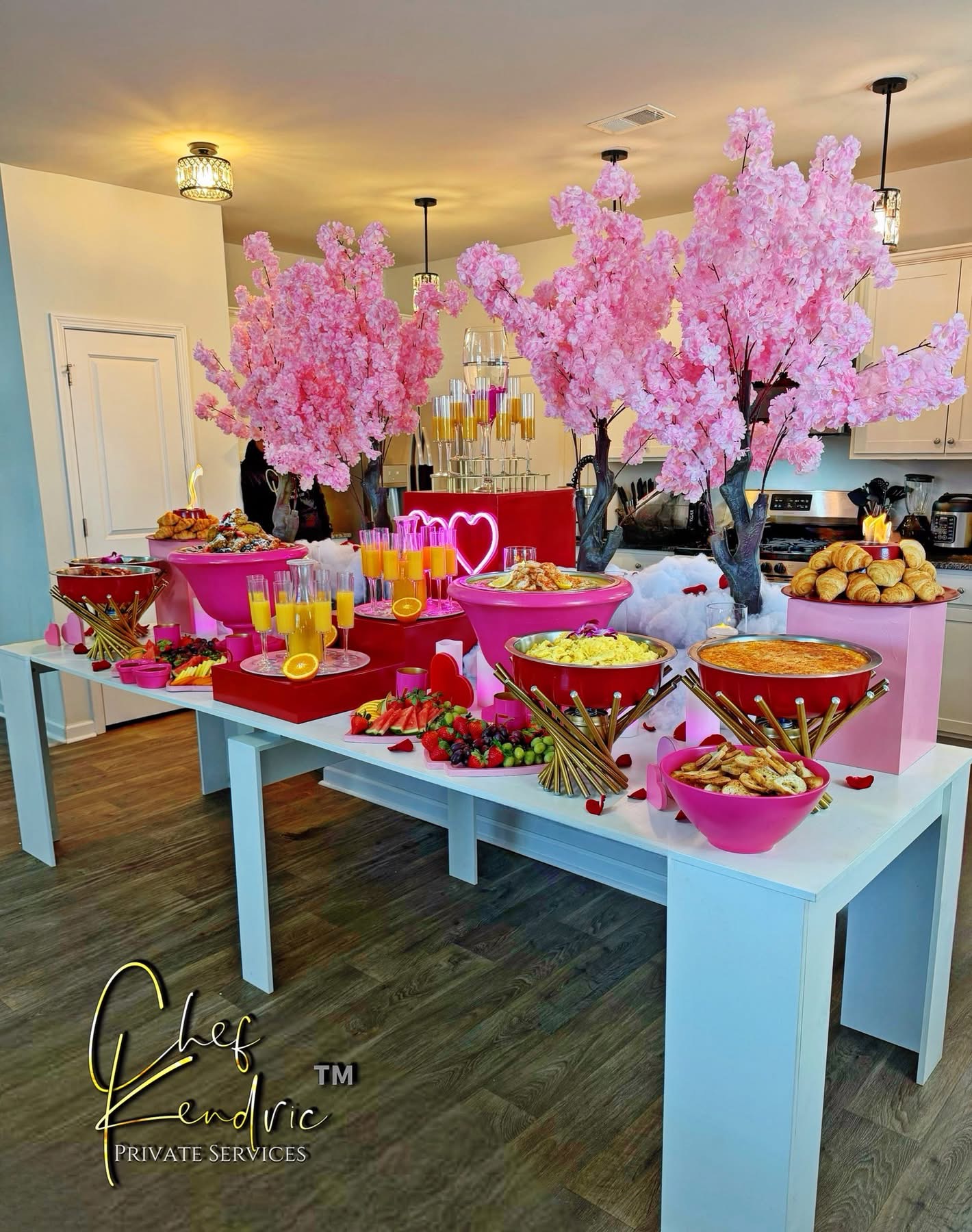Table with pink cherry blossom trees, bowls of snacks, drinks, and decorations in a bright kitchen, celebrating a festive event.
