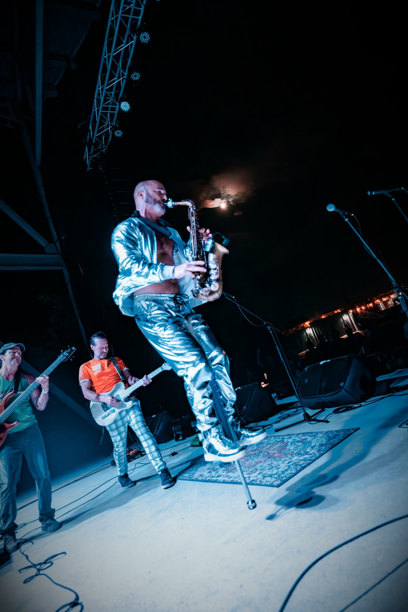 Musicians perform on stage, with one playing a saxophone and others playing guitars, under stage lighting.