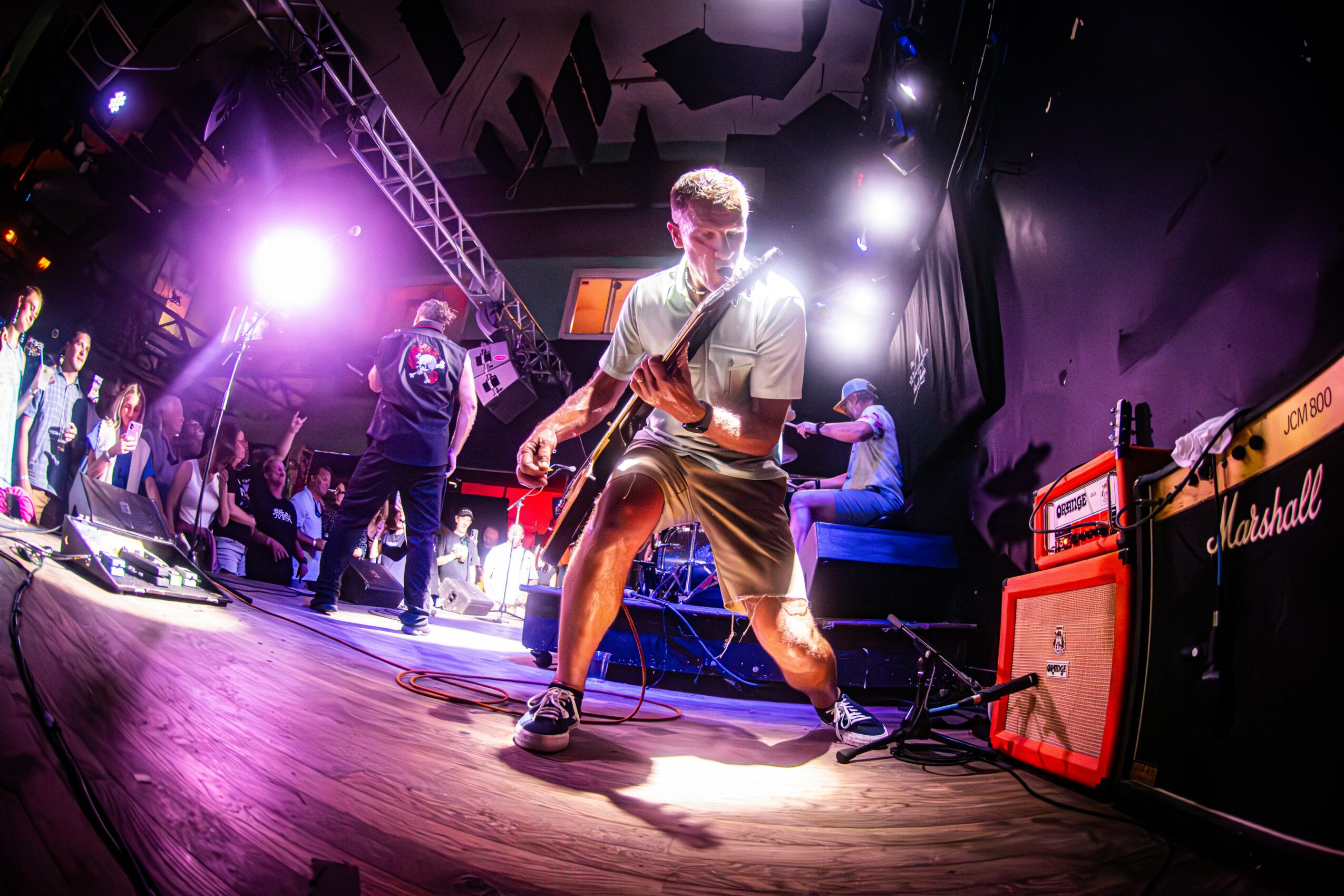 Person playing guitar on stage with bright lights and Marshall amplifier, audience in background, energetic atmosphere.