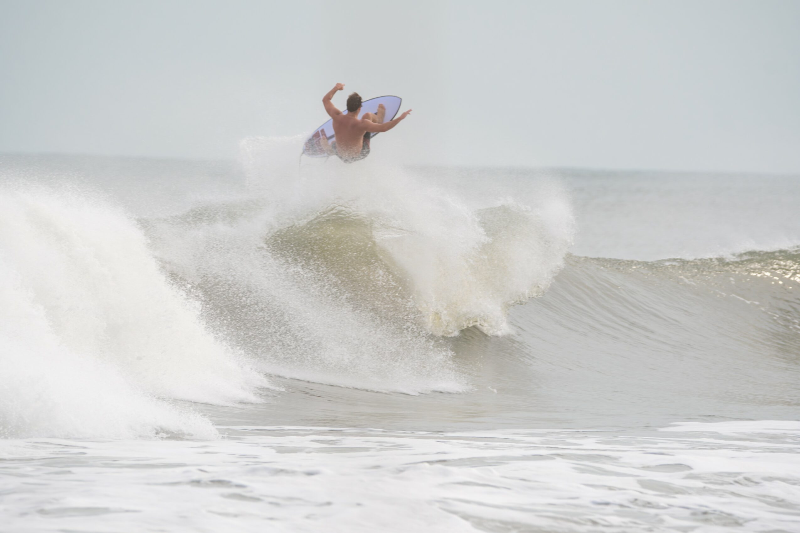 Person surfing on a wave with a clear sky background, holding a surfboard, mid-air above the water, with spray around.