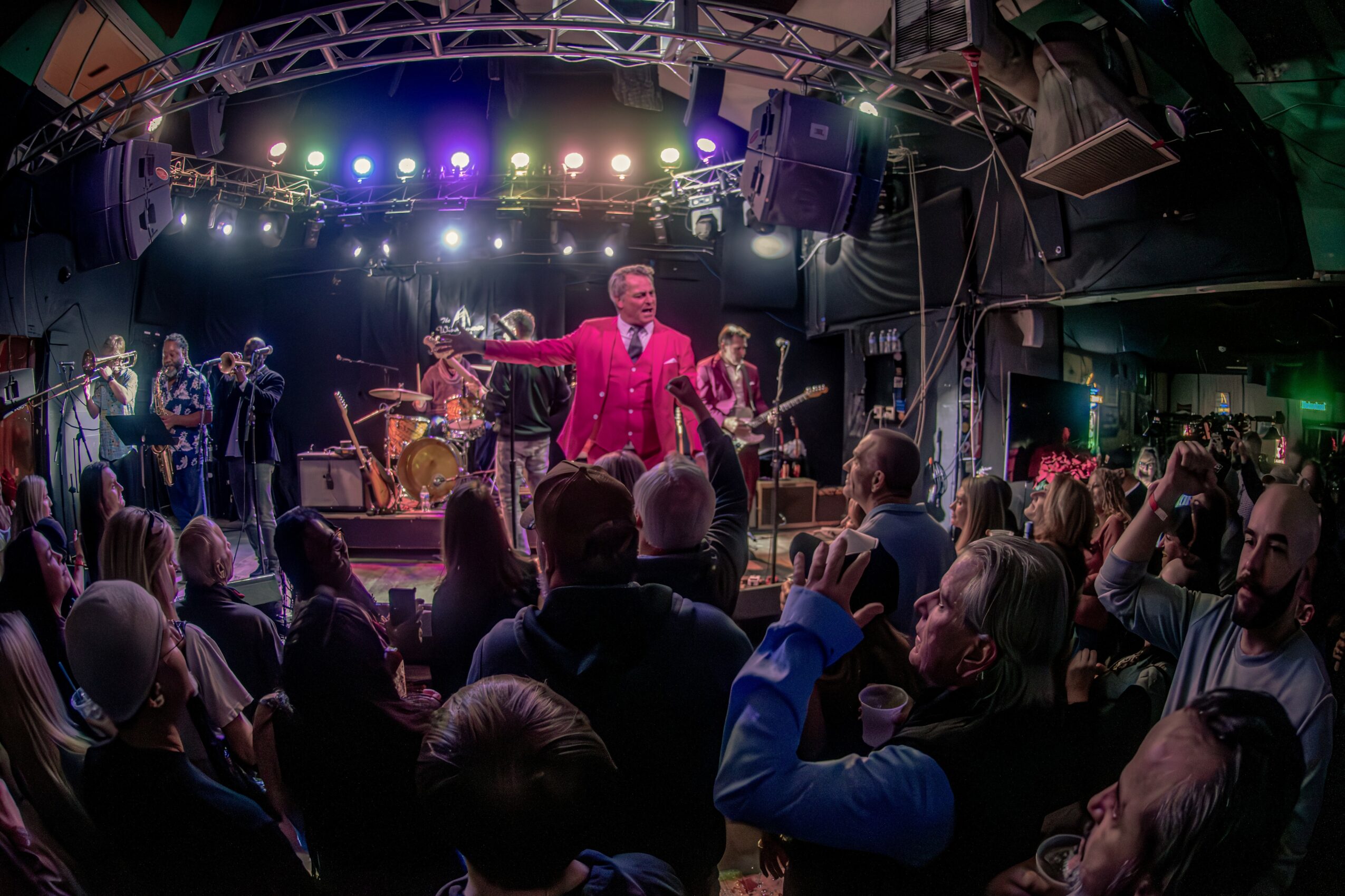 Performer in a pink suit on stage with a band, audience watching in a dimly lit venue with colorful lights.
