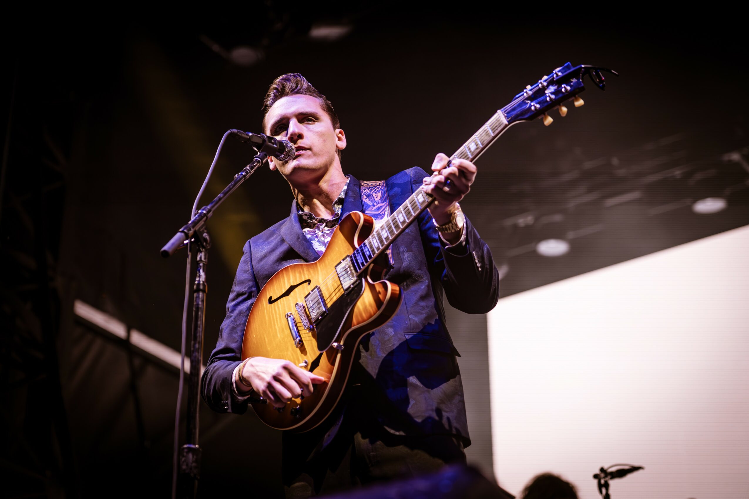 Male musician playing guitar and singing into microphone on stage, with dark background and stage lighting.