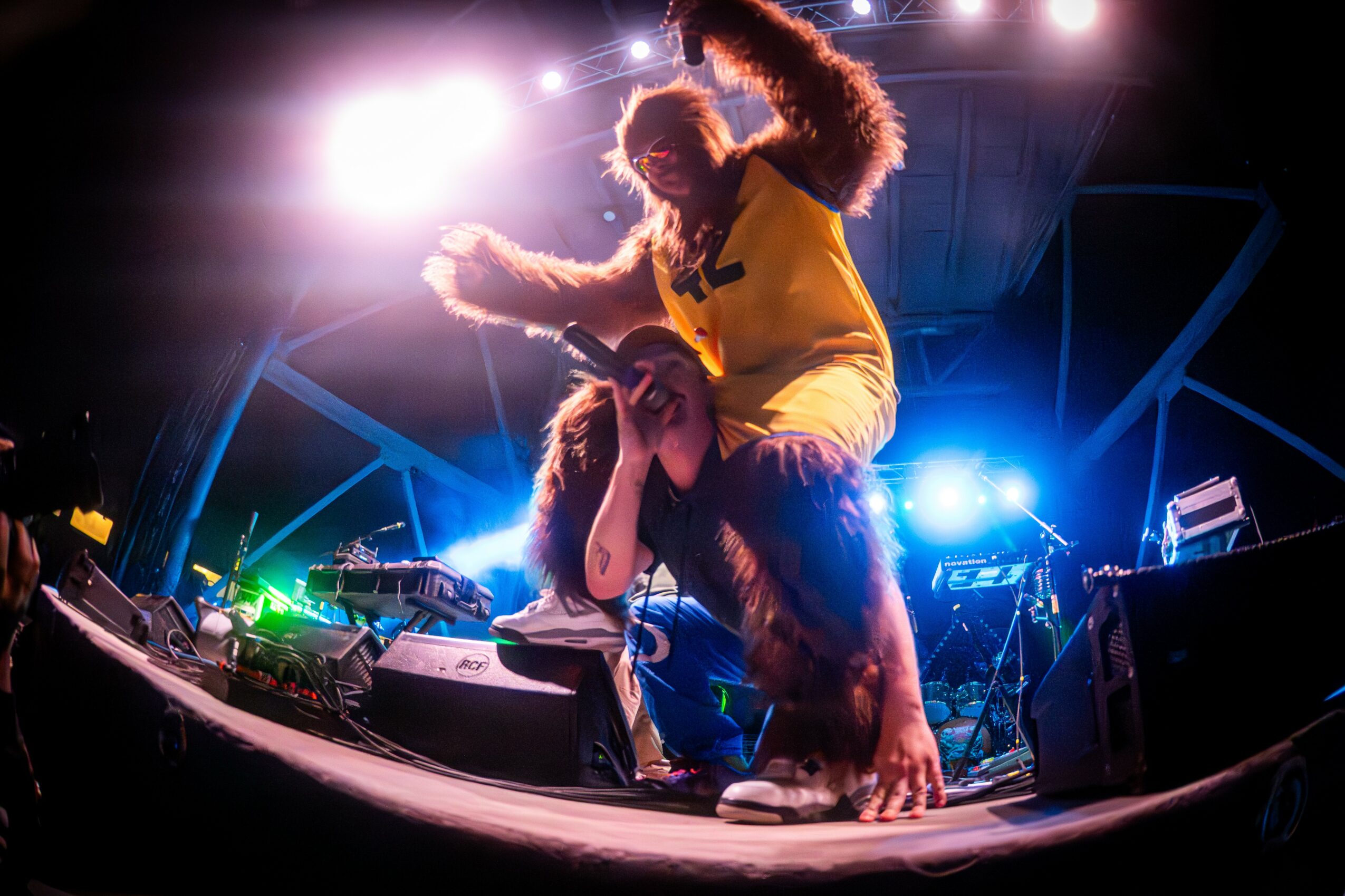 Two people on stage with DJ equipment, one crouching and the other standing, under bright stage lights, in a concert setting.