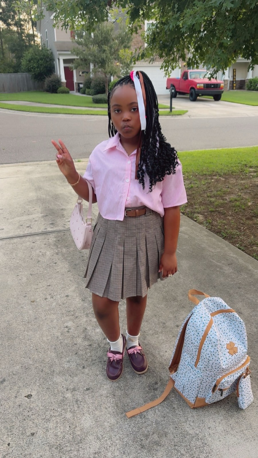 Young girl standing on sidewalk with backpack nearby, wearing pink shirt, gray pleated skirt, and purple shoes.