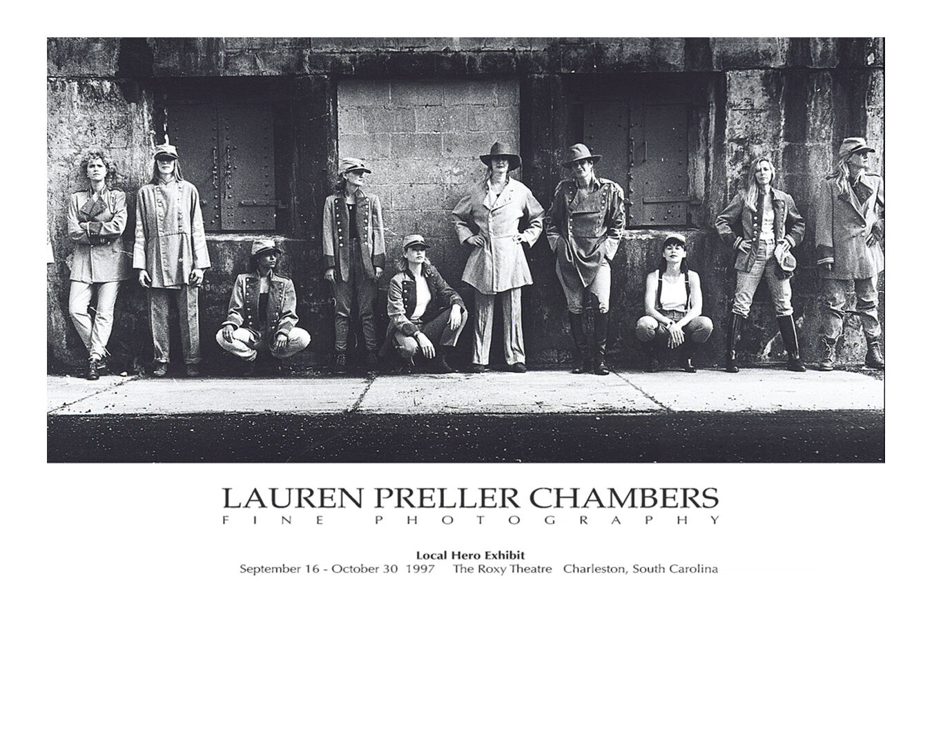 Group of people standing and sitting against a wall, some wearing hats, in black and white photo.