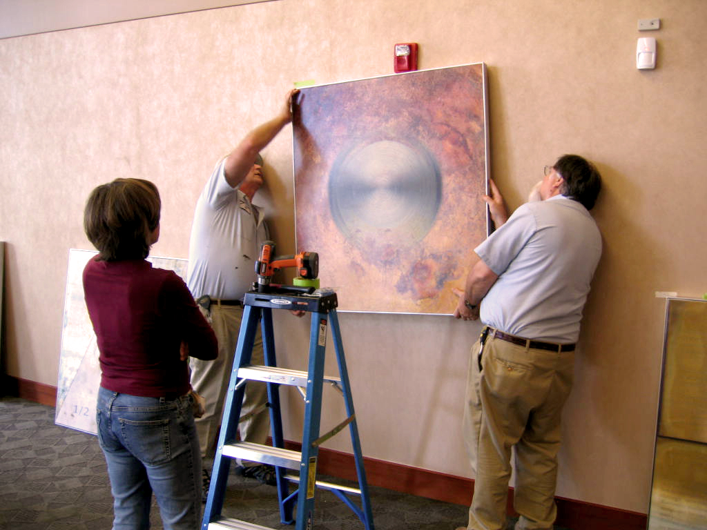 Three people hang a large abstract painting on a beige wall, with a blue ladder nearby. One person holds the painting, another adjusts it, and a third observes.