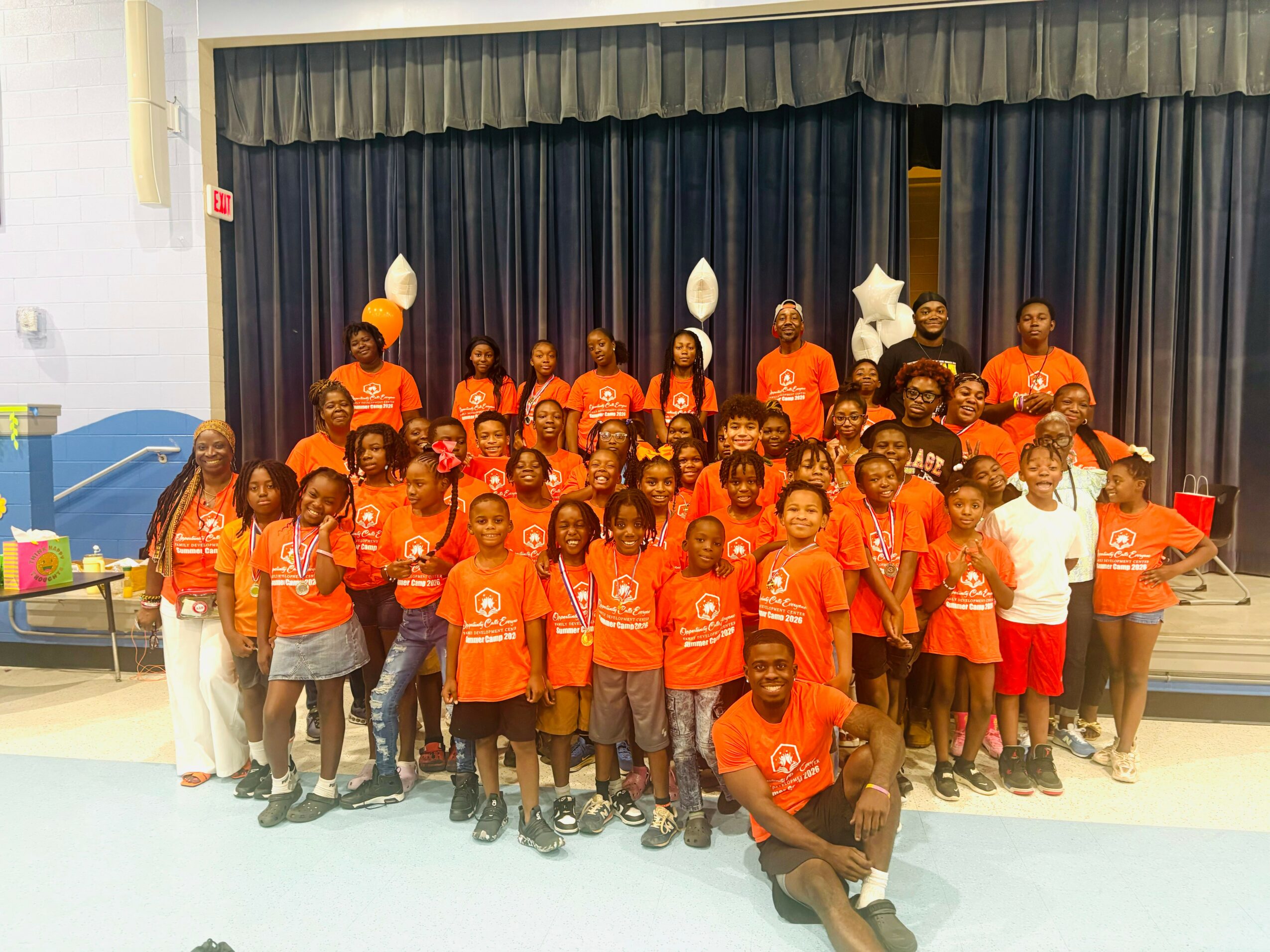 Group of children and adults on stage, wearing matching orange shirts, with balloons and a curtain backdrop.