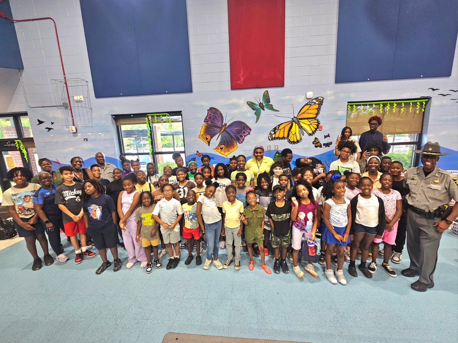 Group of children and adults standing indoors in front of a wall with butterfly decorations and a flag, smiling at the camera.