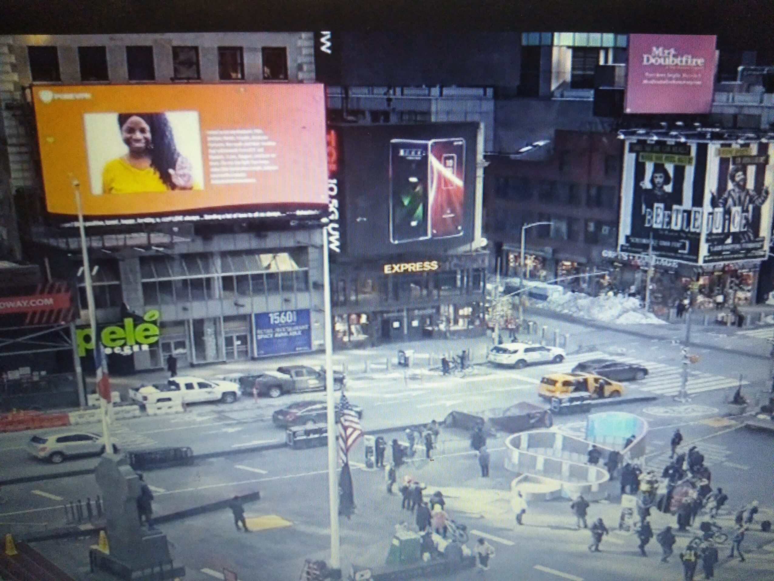 City street scene at night with billboards, cars, and pedestrians crossing the road.