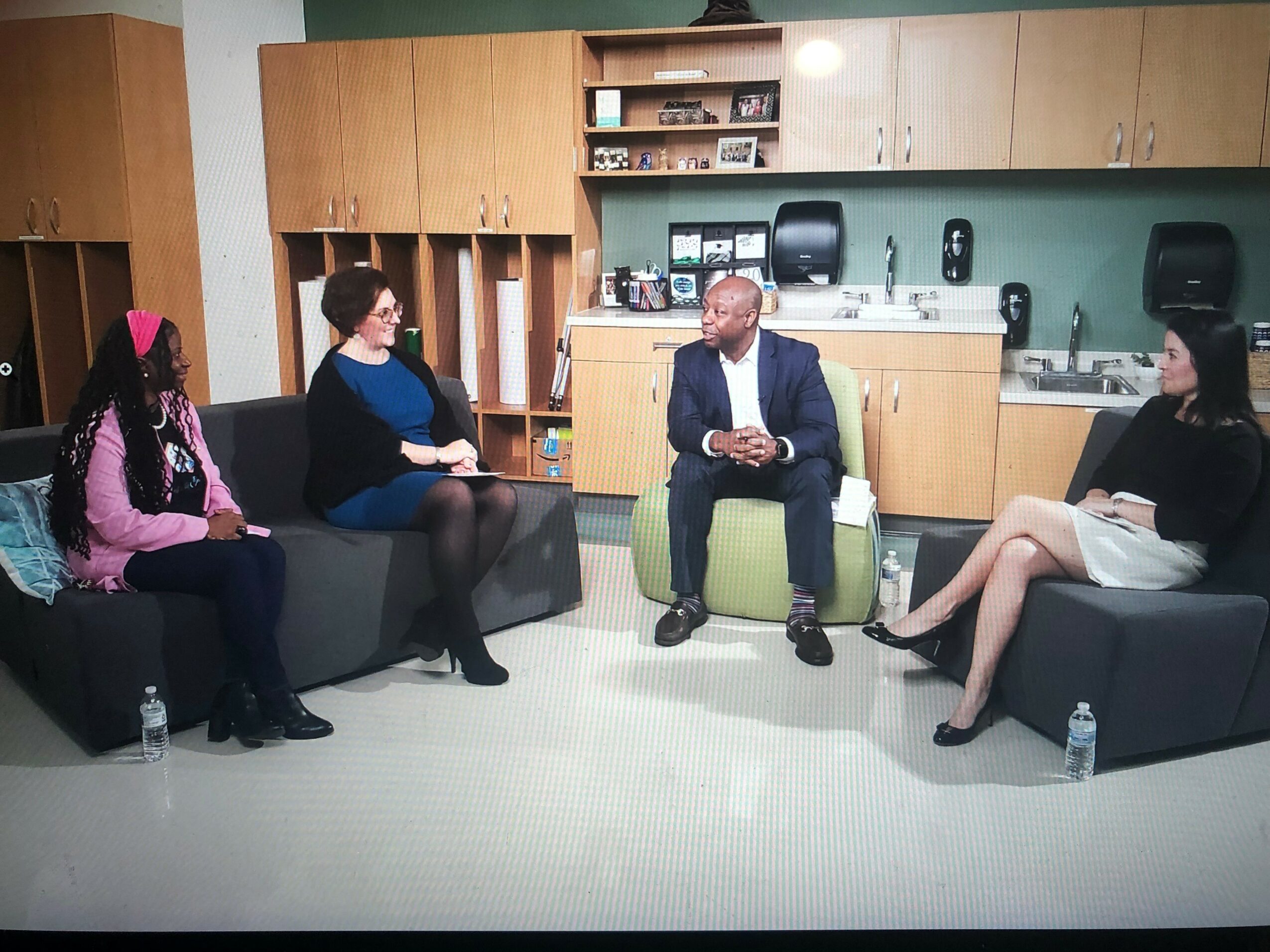 Four people seated in a room with wooden cabinets, discussing, with water bottles on the floor, and a green wall behind them.