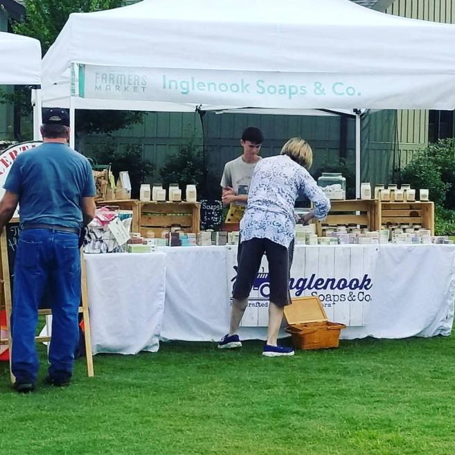 Person at outdoor soap booth with table and products, others nearby, tent with signage, grassy area, building in background.
