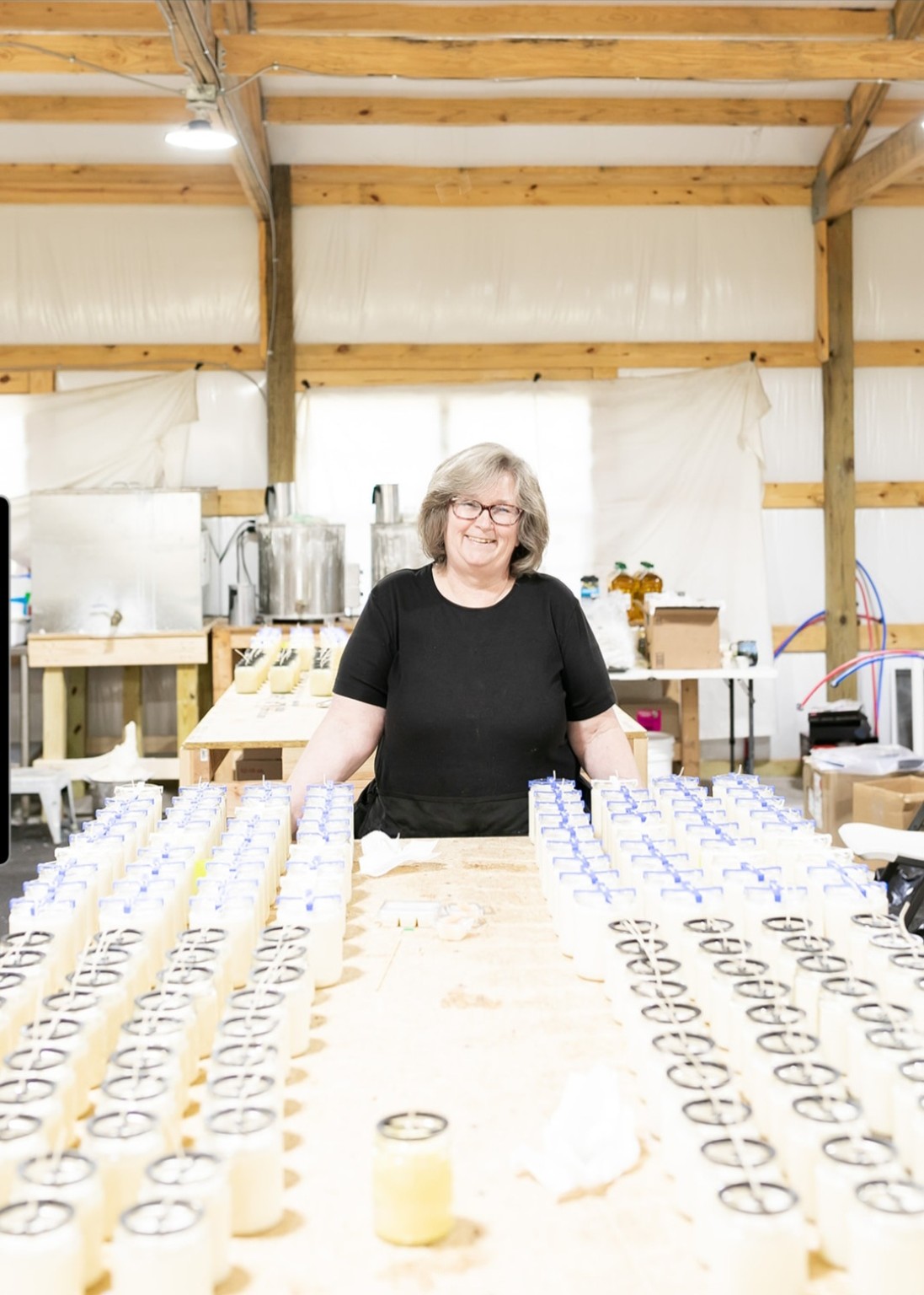 Woman with glasses and black shirt standing behind a table with rows of jewelry, in a workshop with wooden beams.