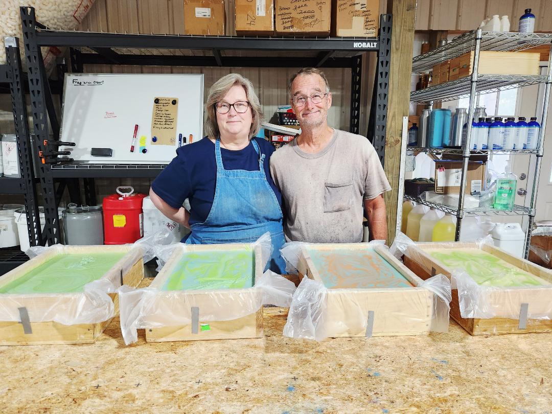 Two people standing behind a table with containers covered in plastic, in a storage room with shelves and boxes.