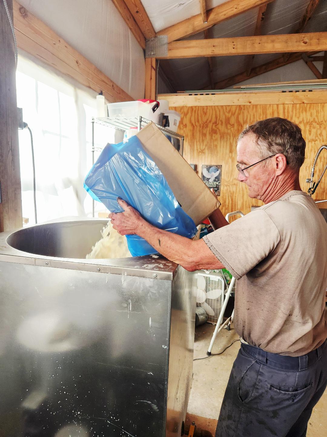 Man pouring blue bag contents into large metal container in workshop with wooden walls and ceiling