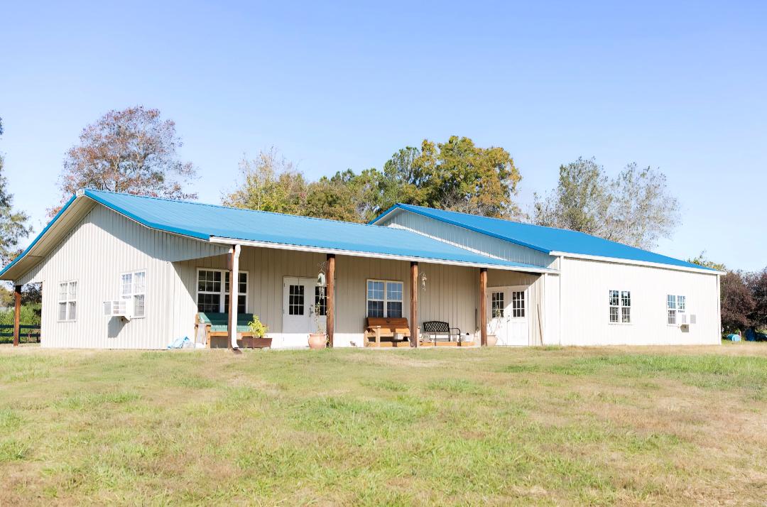Single-story building with a blue roof, white walls, and a porch, set in a grassy area with trees in the background.