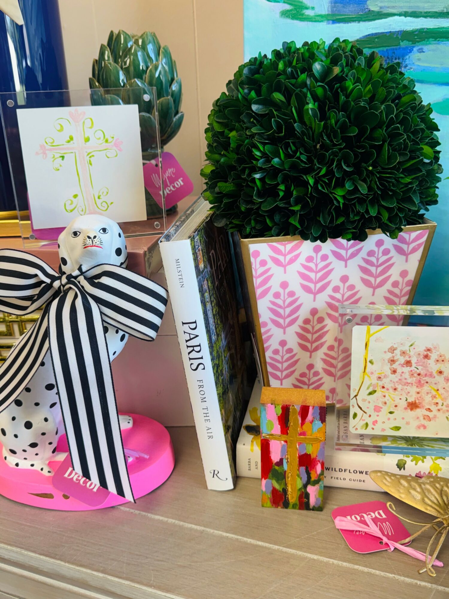 Decorative plants, books, and colorful items on a table, with a pink and white patterned container and a striped bow.