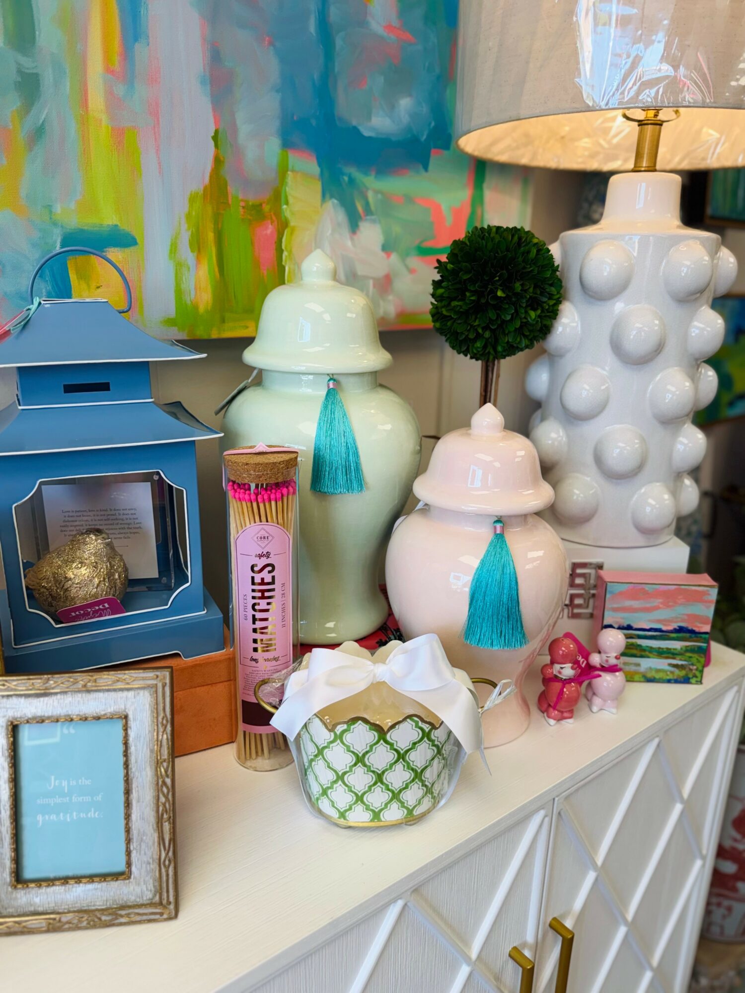 Decorative items on a white surface include vases, a lamp, a small tree, a pink toy, and a framed picture, with colorful abstract background.