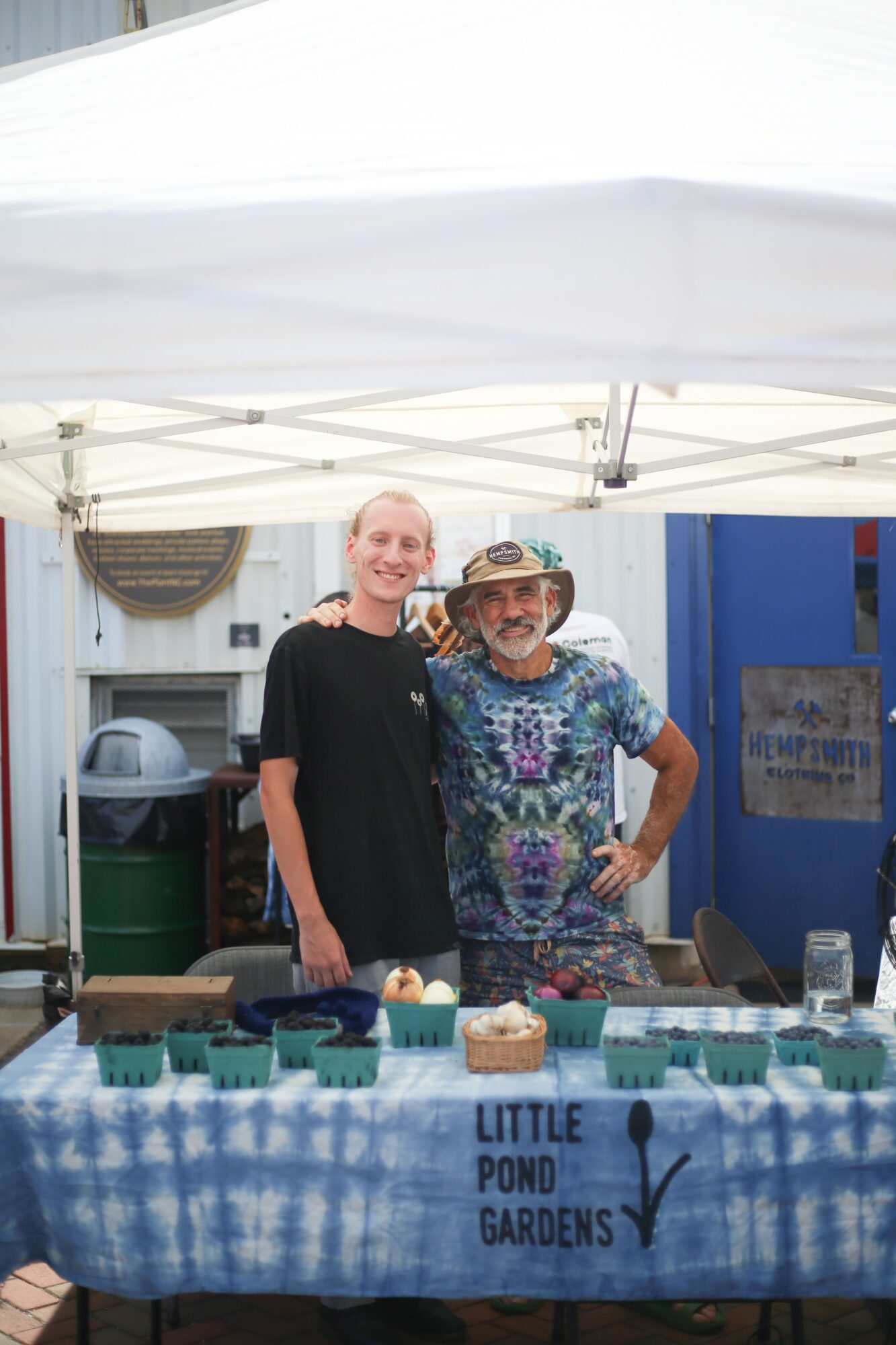 Two people standing behind a table with small potted plants, under a white canopy, in front of a blue building.