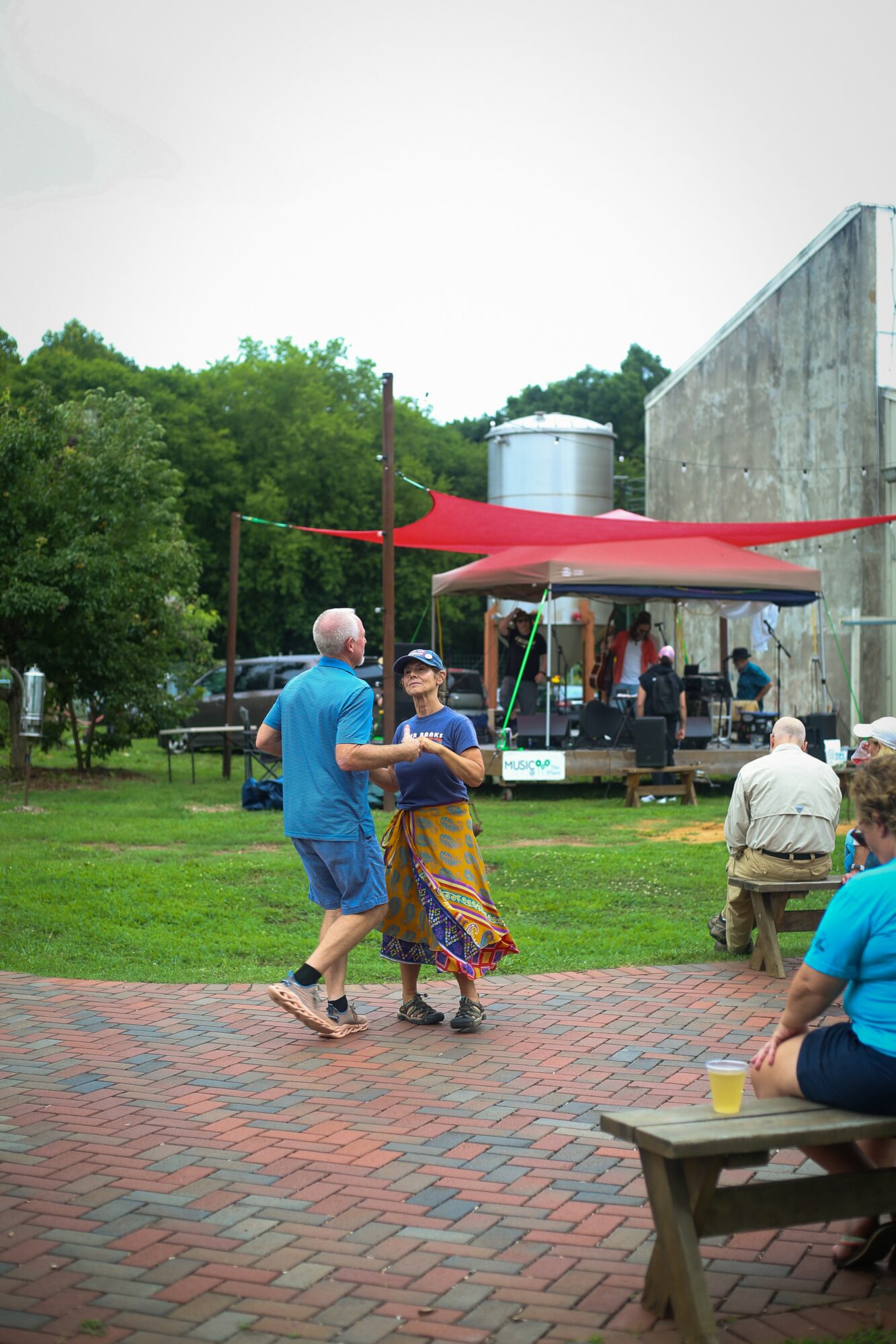 Two people dancing outdoors on a brick path, with a stage, red canopy, and trees in the background.