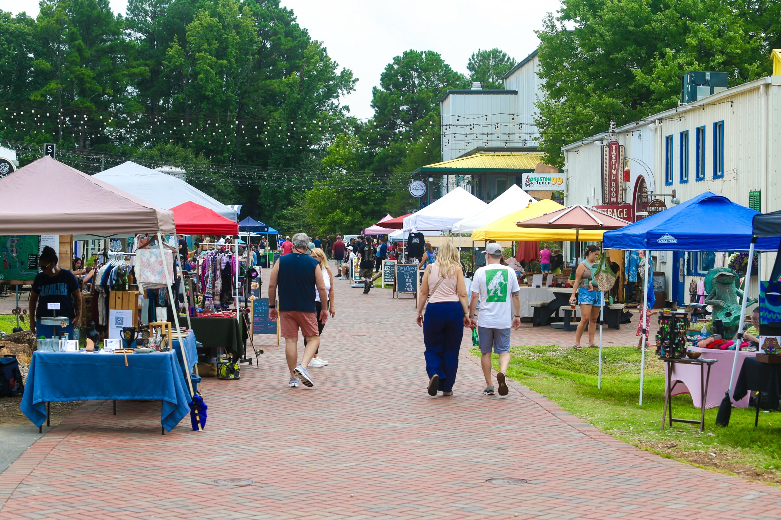 Outdoor market with tents and people walking on a brick path, trees and buildings in the background.