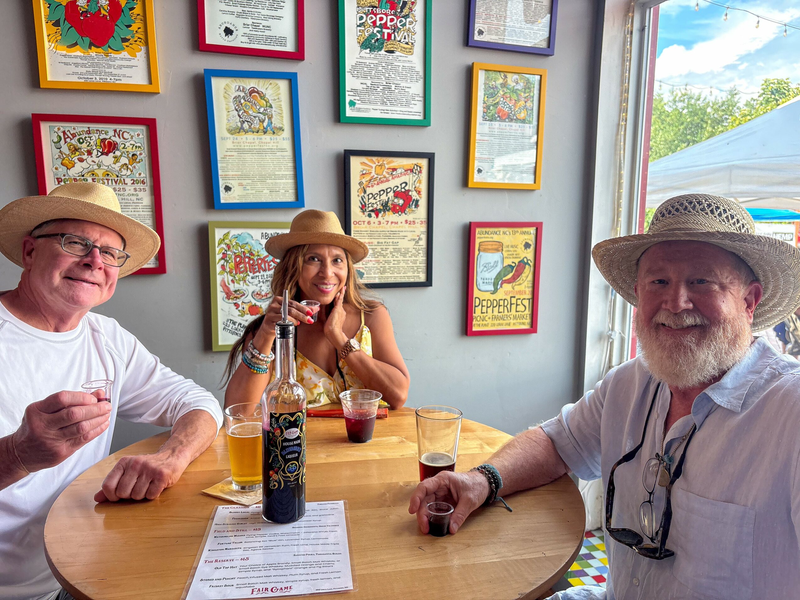 Three people sitting at a table inside a room with framed artwork on the wall, enjoying drinks and smiling.