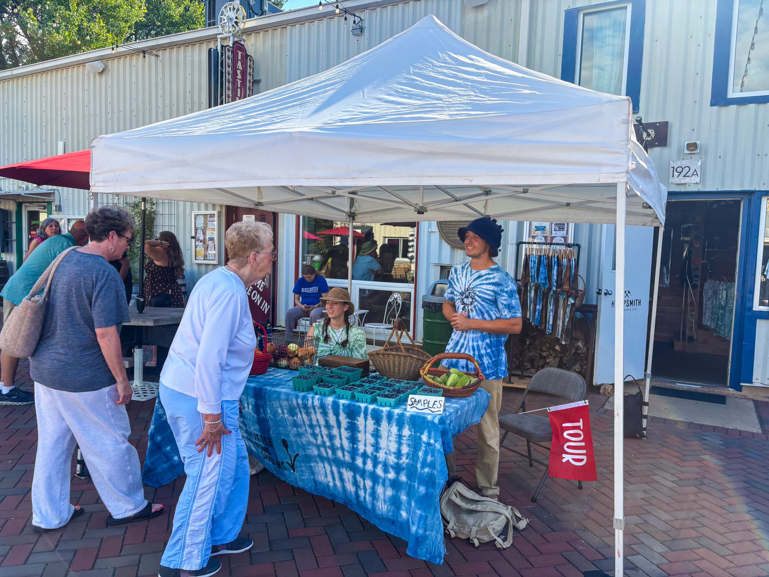 People gather around a market stall with a blue tablecloth under a white canopy, selling jewelry and accessories outdoors.