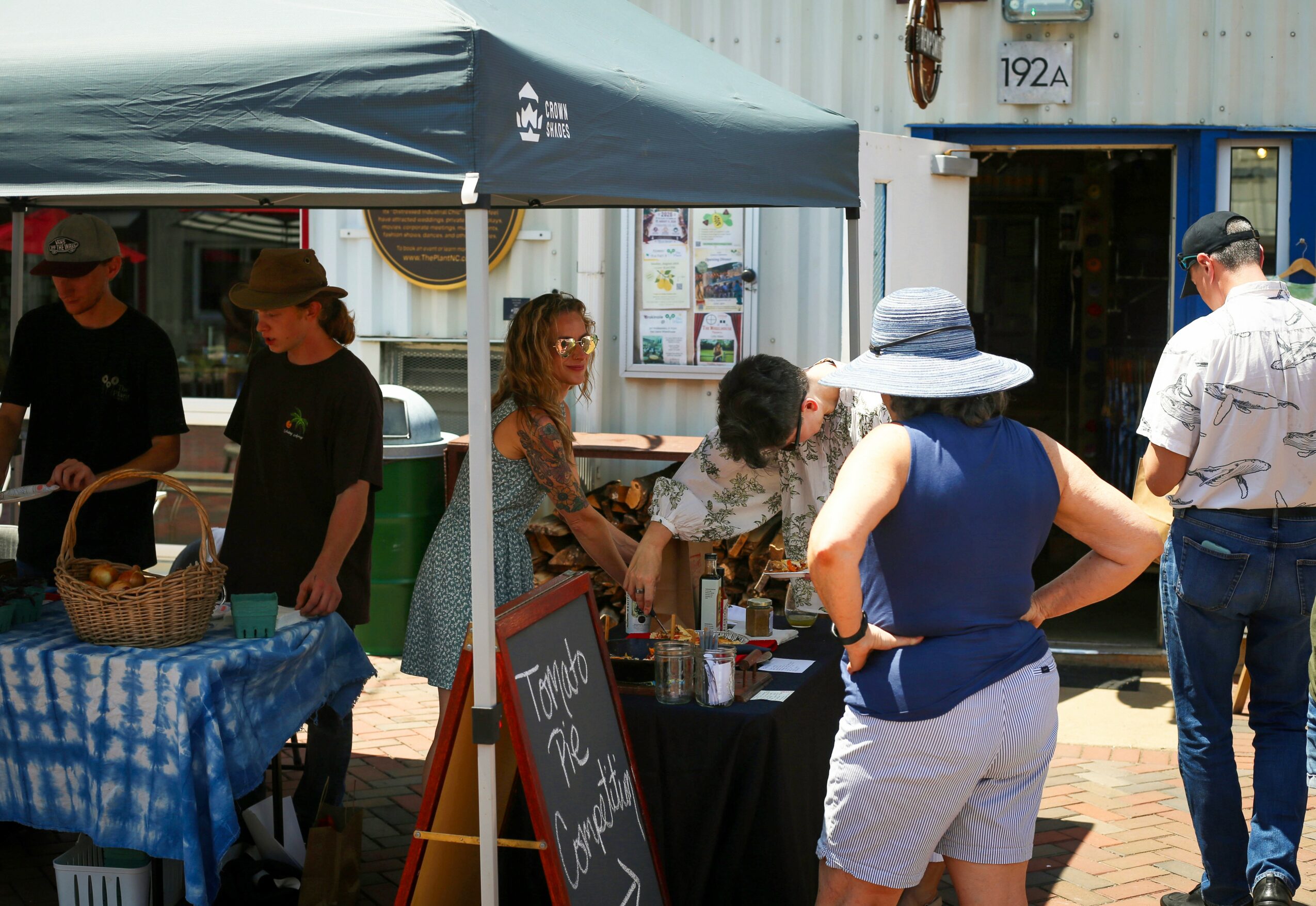 People gather outside a shop under a canopy, some browsing tables with items, others talking, with a chalkboard sign nearby.