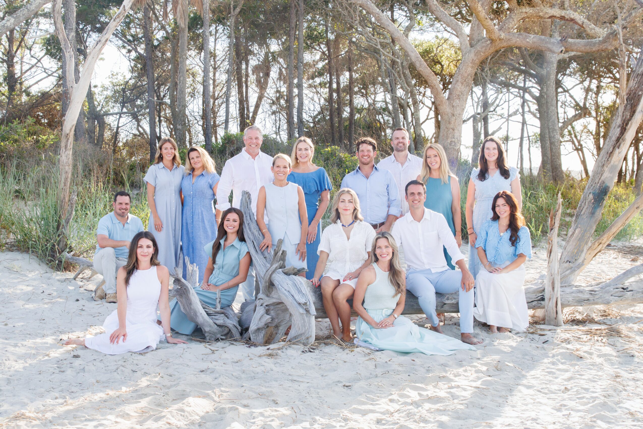 Group of people outdoors on sandy ground with trees, some sitting and some standing, dressed in light-colored clothing.