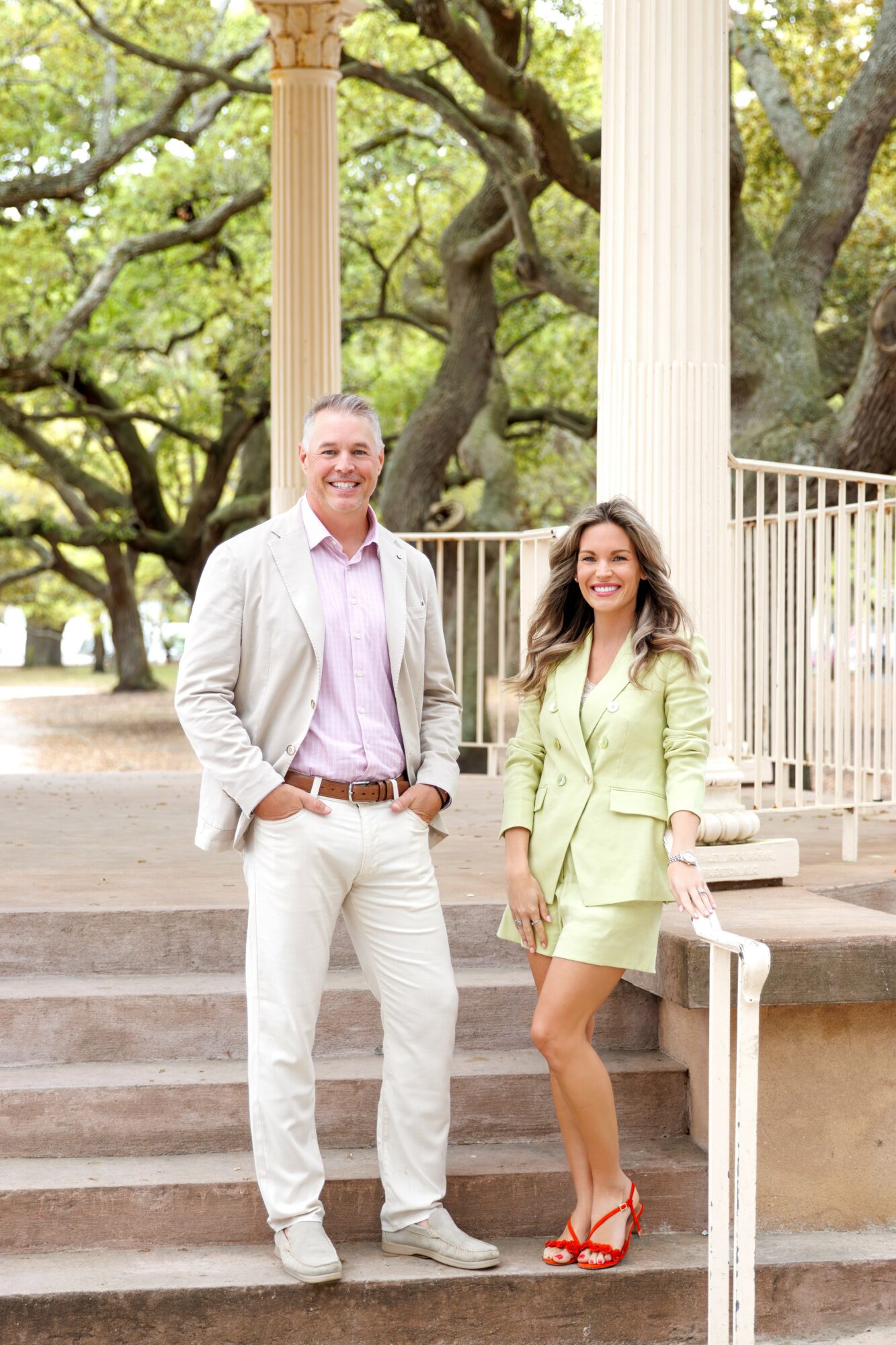 Two people, a man and a woman, stand on steps outdoors with trees and columns in background.