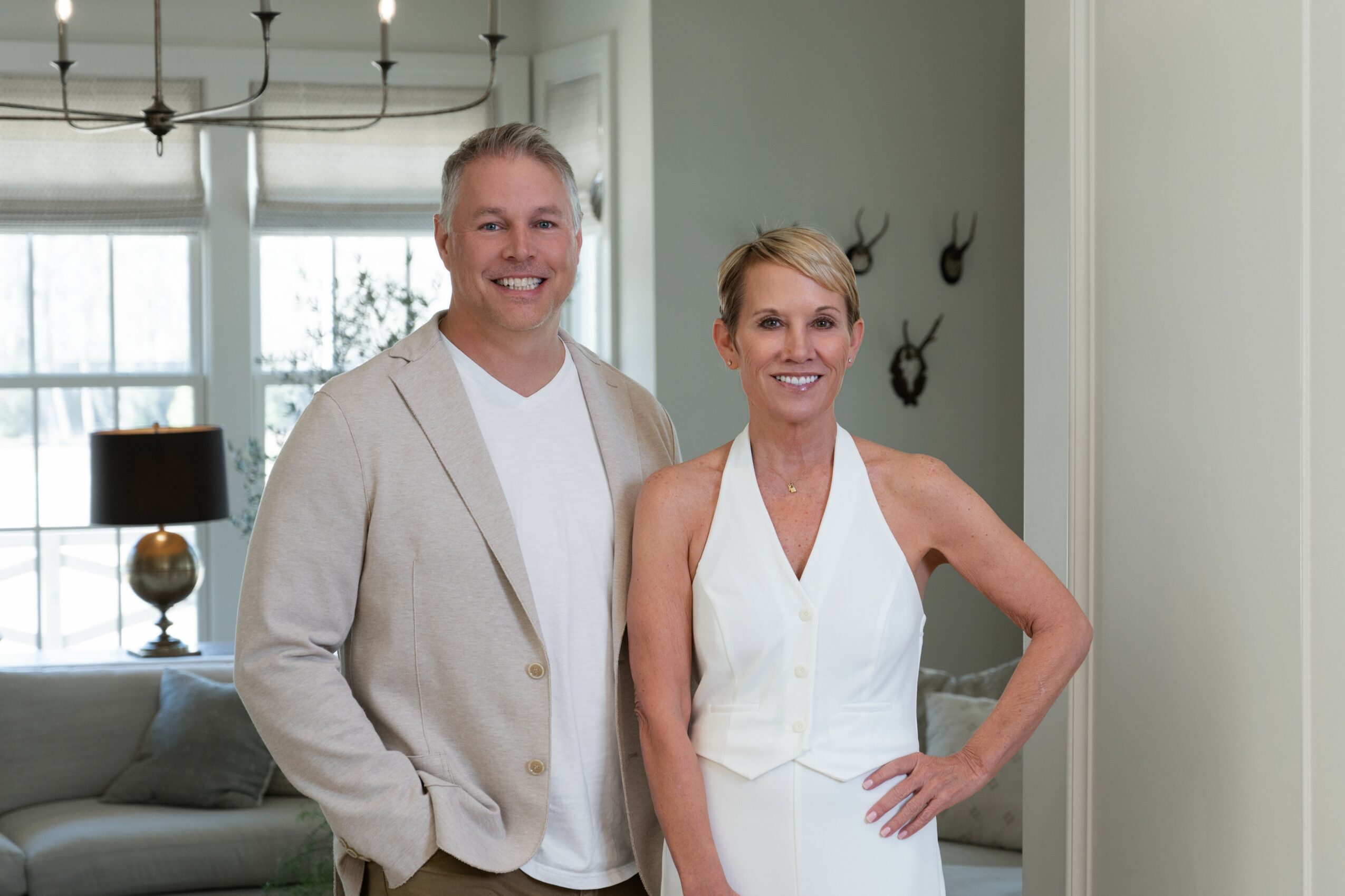 Two women standing indoors, smiling, with a living room background including a window, lamp, and wall decorations.