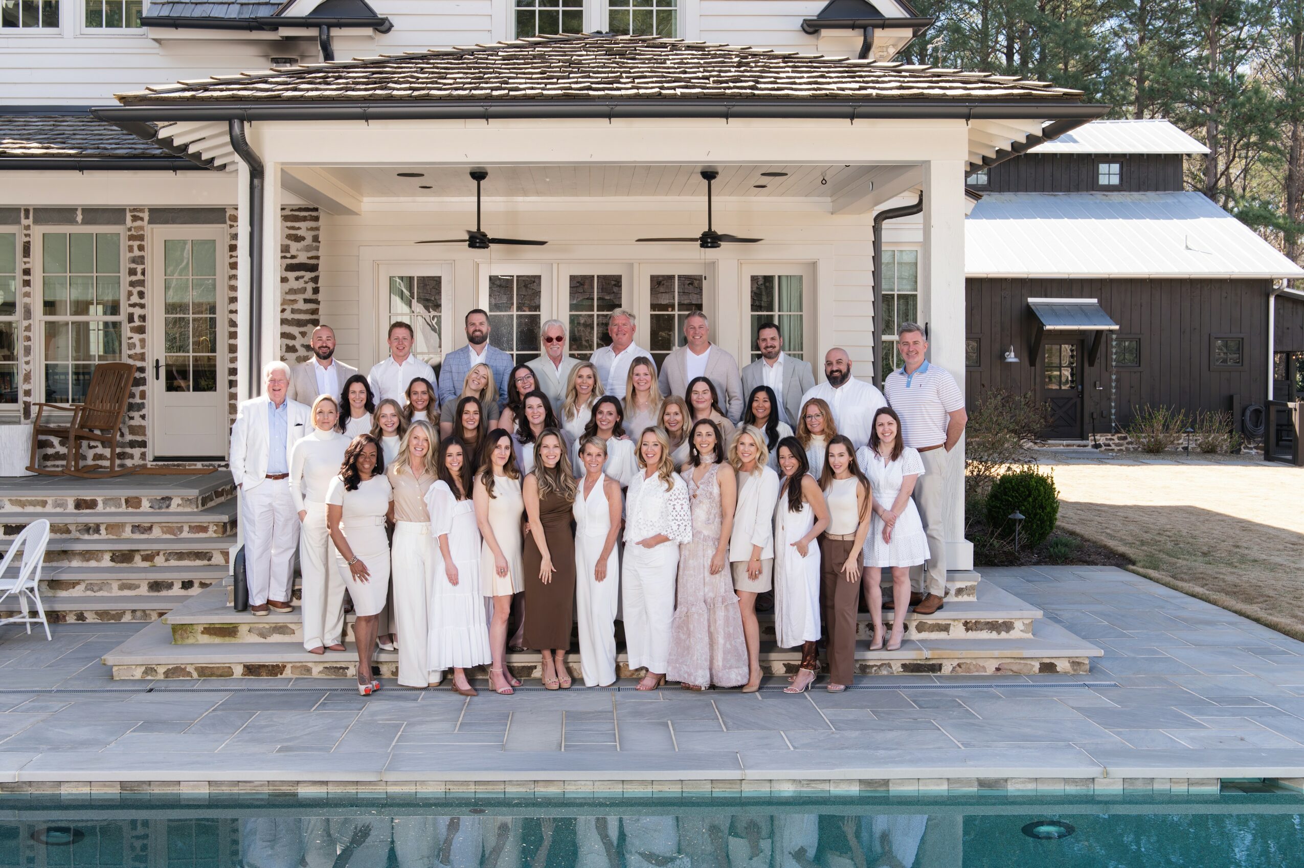 Group of people in white clothing standing on steps outside a house with a porch, near a pool.