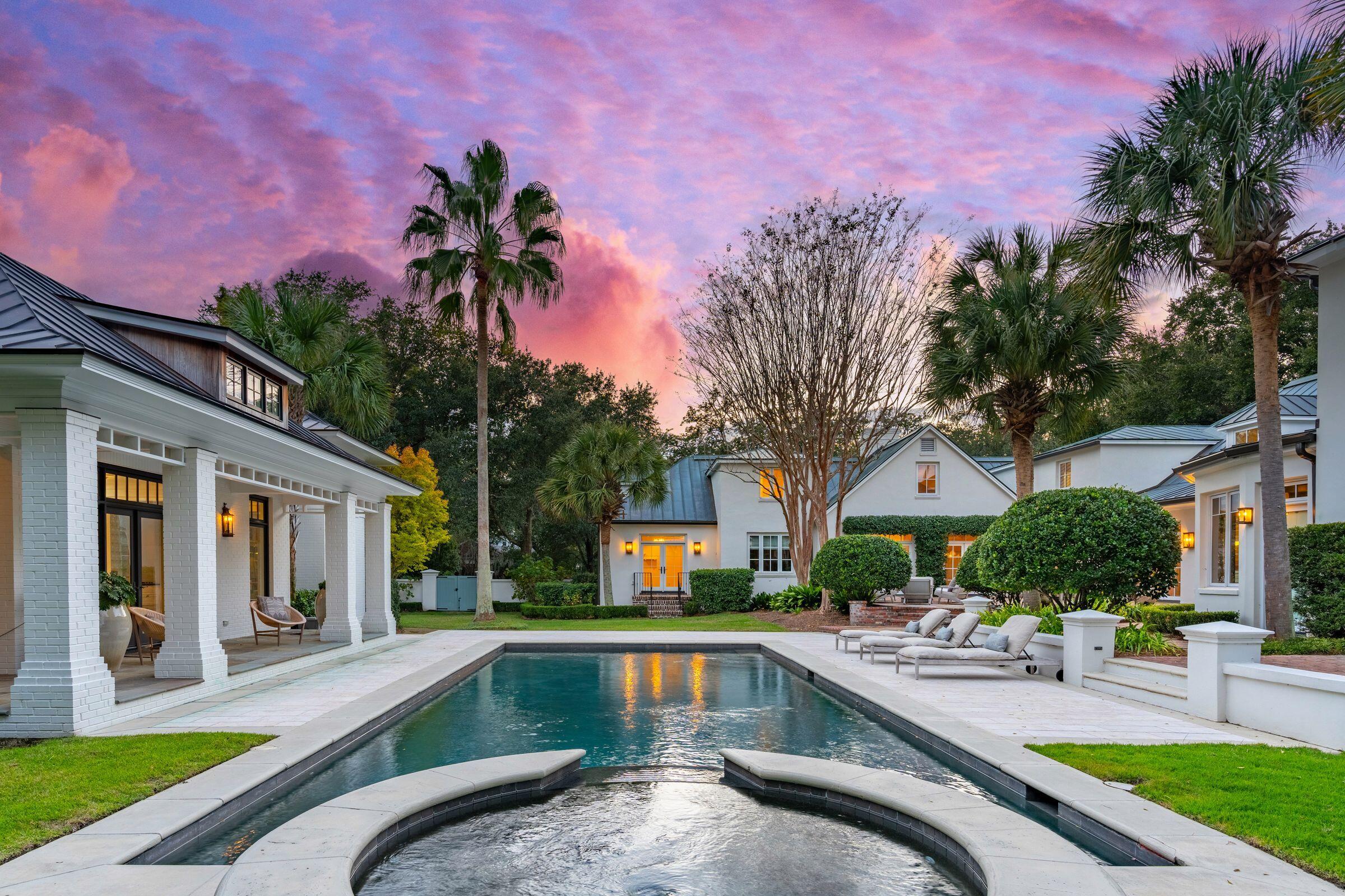 Swimming pool in a backyard with lounge chairs, palm trees, and houses under a colorful sunset sky.