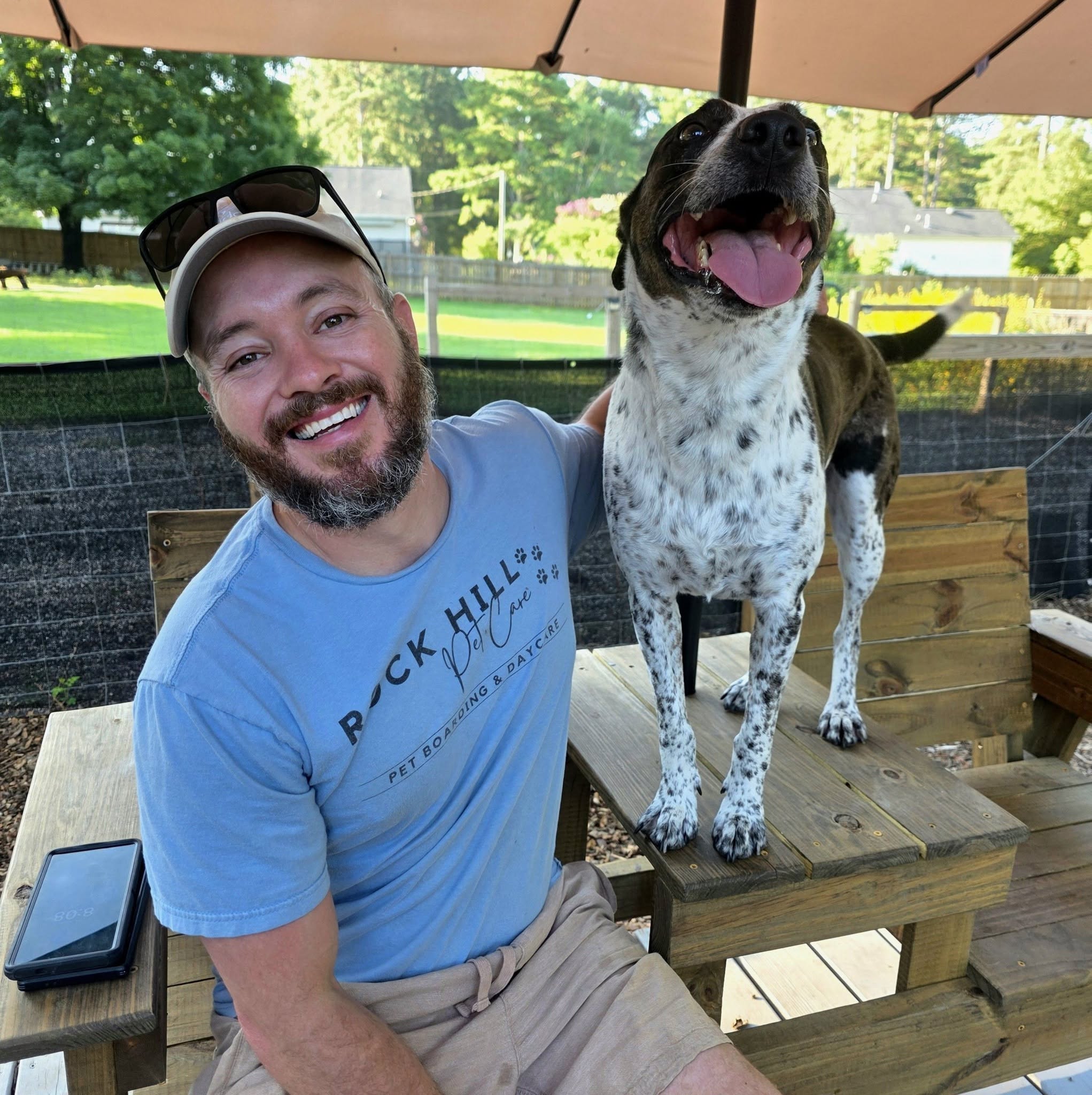 Man with a beard and a cap smiling next to a large dog on a wooden bench outdoors.