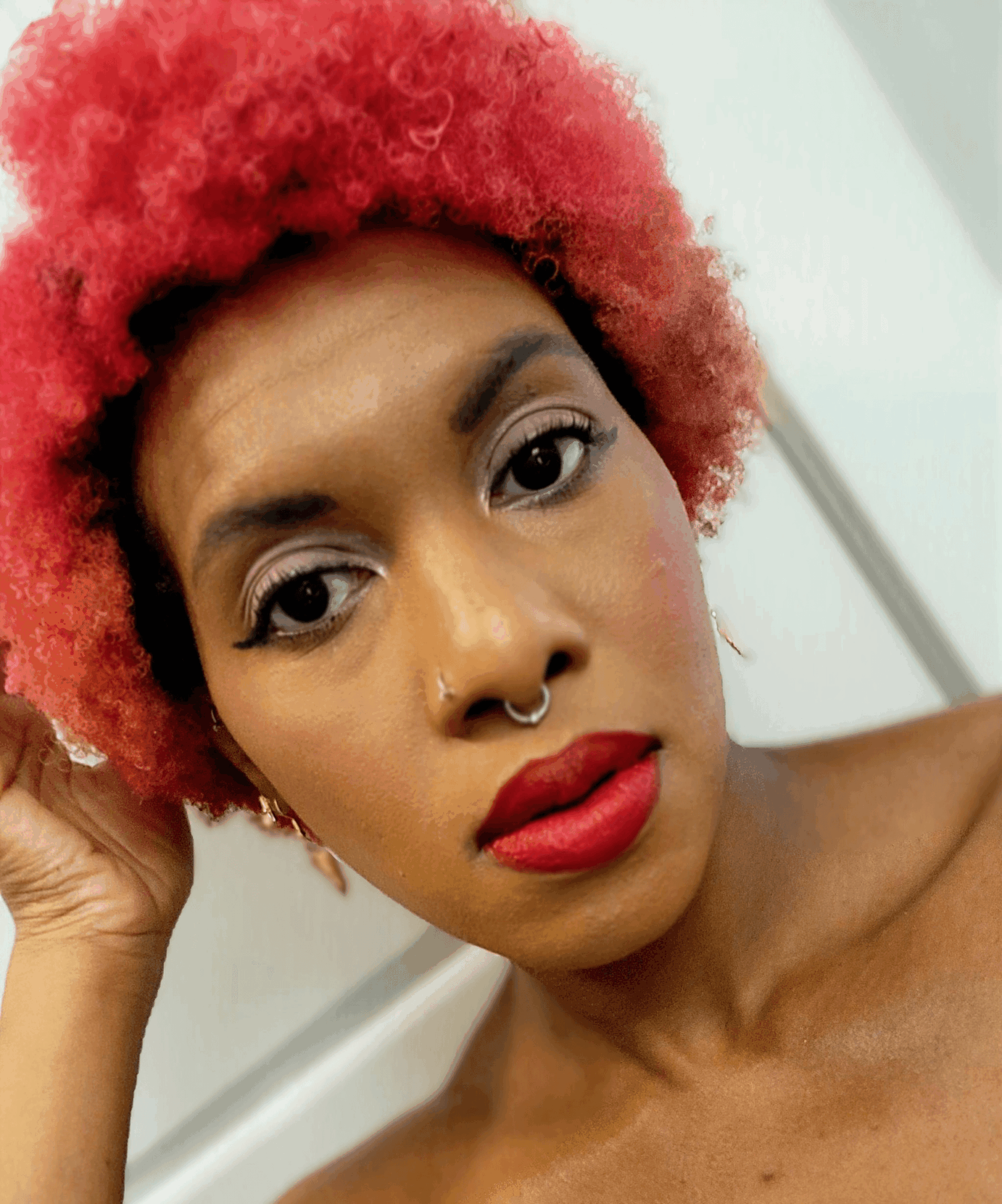 Close-up of a woman with red curly hair, makeup, and a septum piercing, looking at the camera.