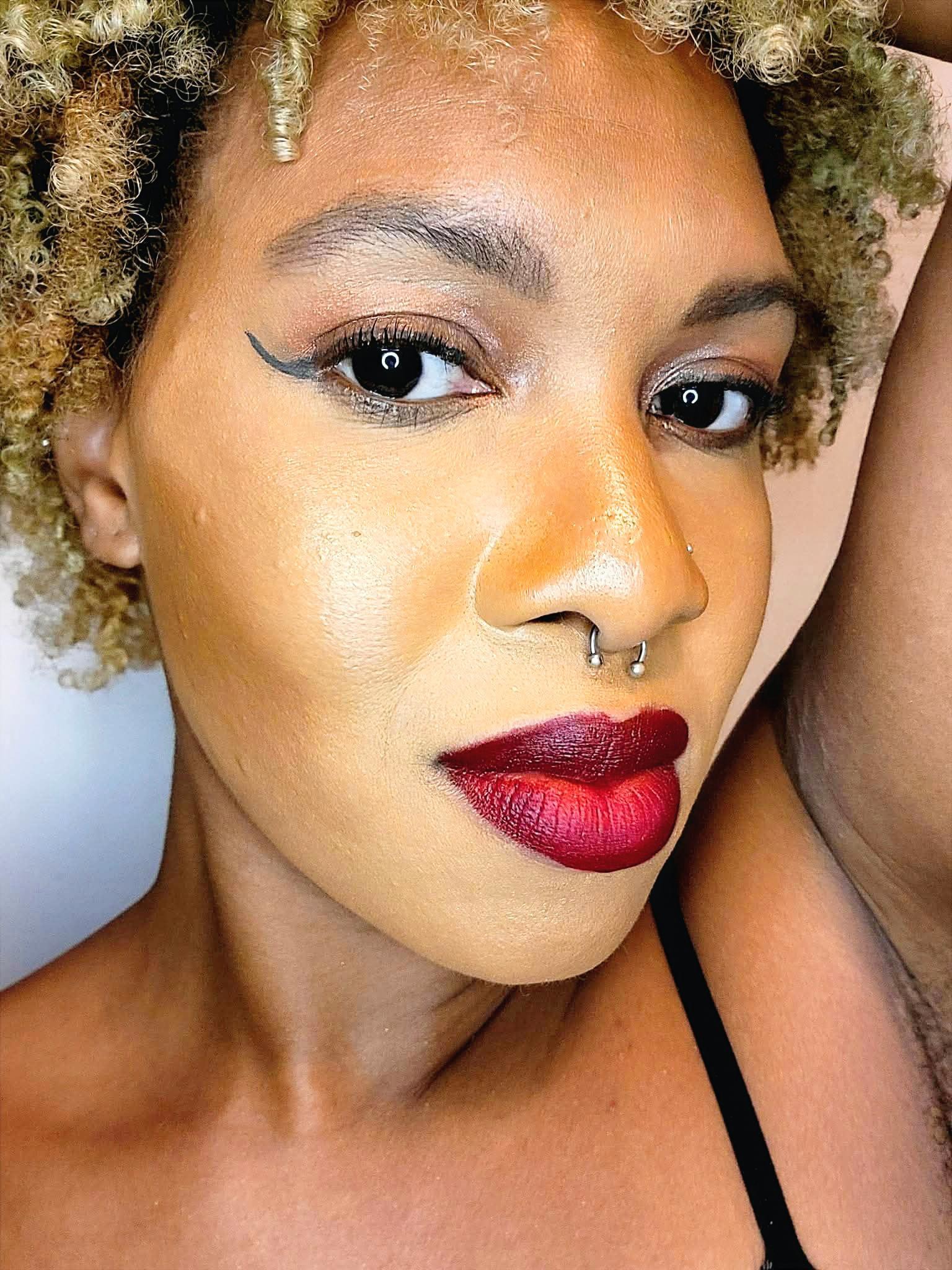 Close-up of a woman with curly hair, bold makeup, and a septum piercing, looking at the camera.