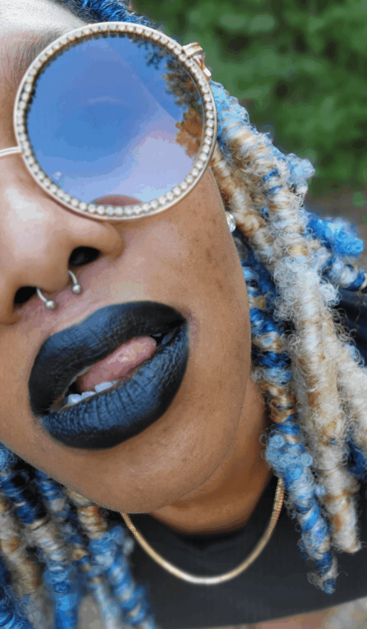 Close-up of a person with sunglasses, black lipstick, and dreadlocks outdoors.