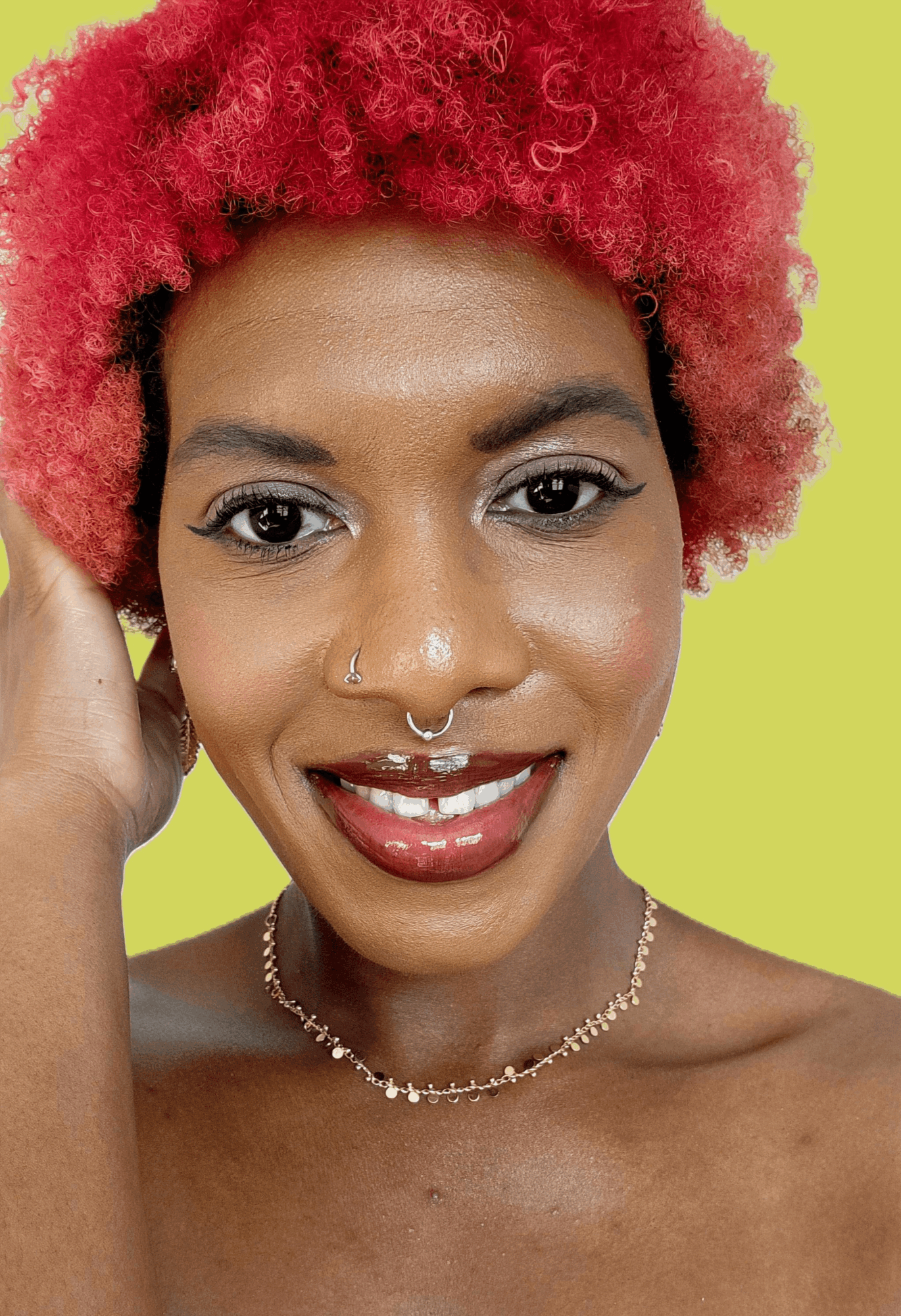 Close-up of a woman with curly red hair, smiling, wearing a necklace and a nose ring, against a yellow background.