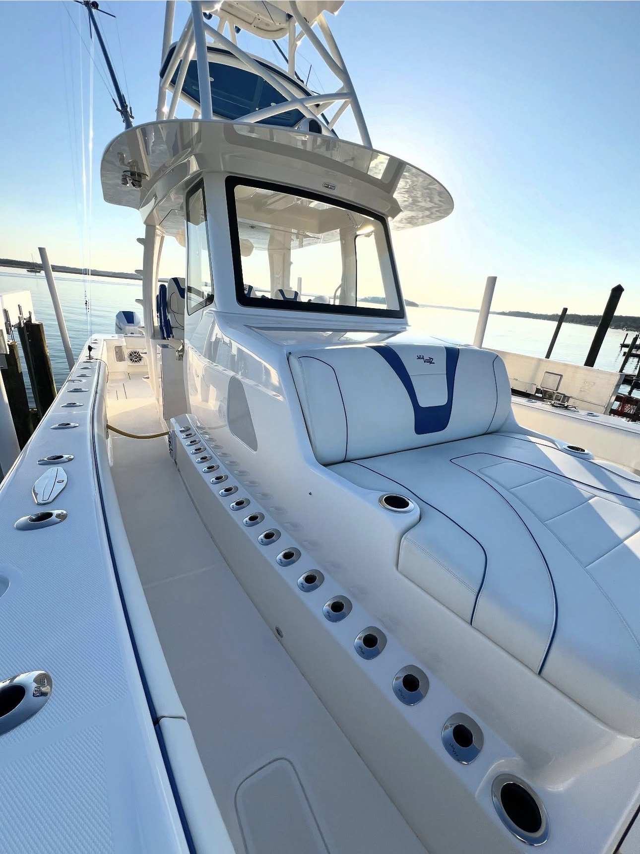 View of a white boat with seating and a steering area, docked at a marina with water and sky in background.