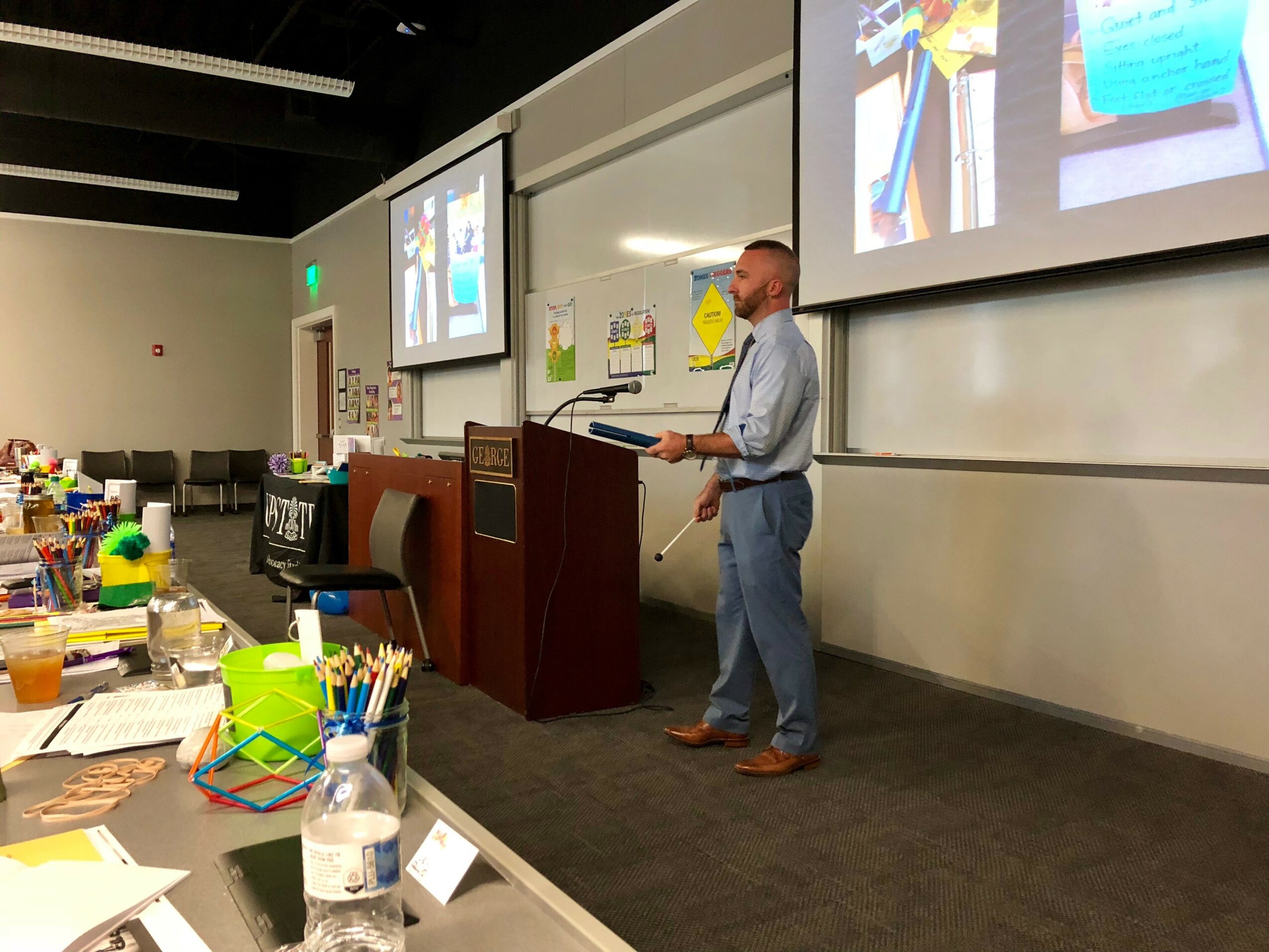 Man standing at podium giving a presentation in a classroom or conference room with screens displaying slides, tables with supplies in foreground.
