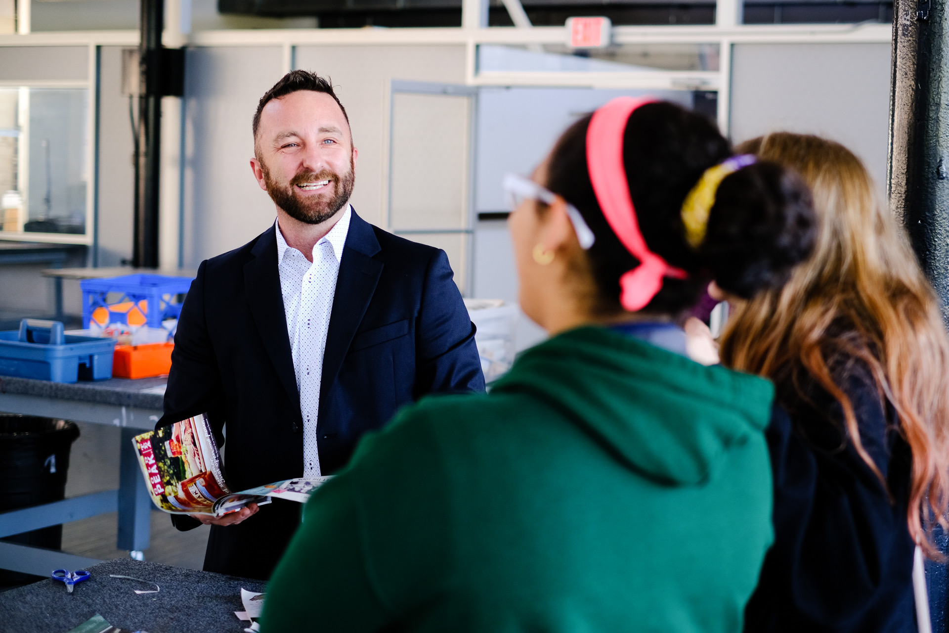 Man in suit smiling and holding a colorful box, talking to two women with their backs to the camera, in an industrial setting.