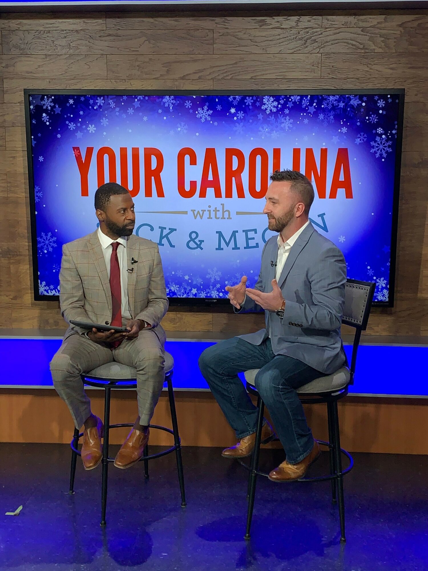 Two men seated on chairs in a television studio, with a large screen behind them displaying 'YOUR CAROLINA' and festive graphics.