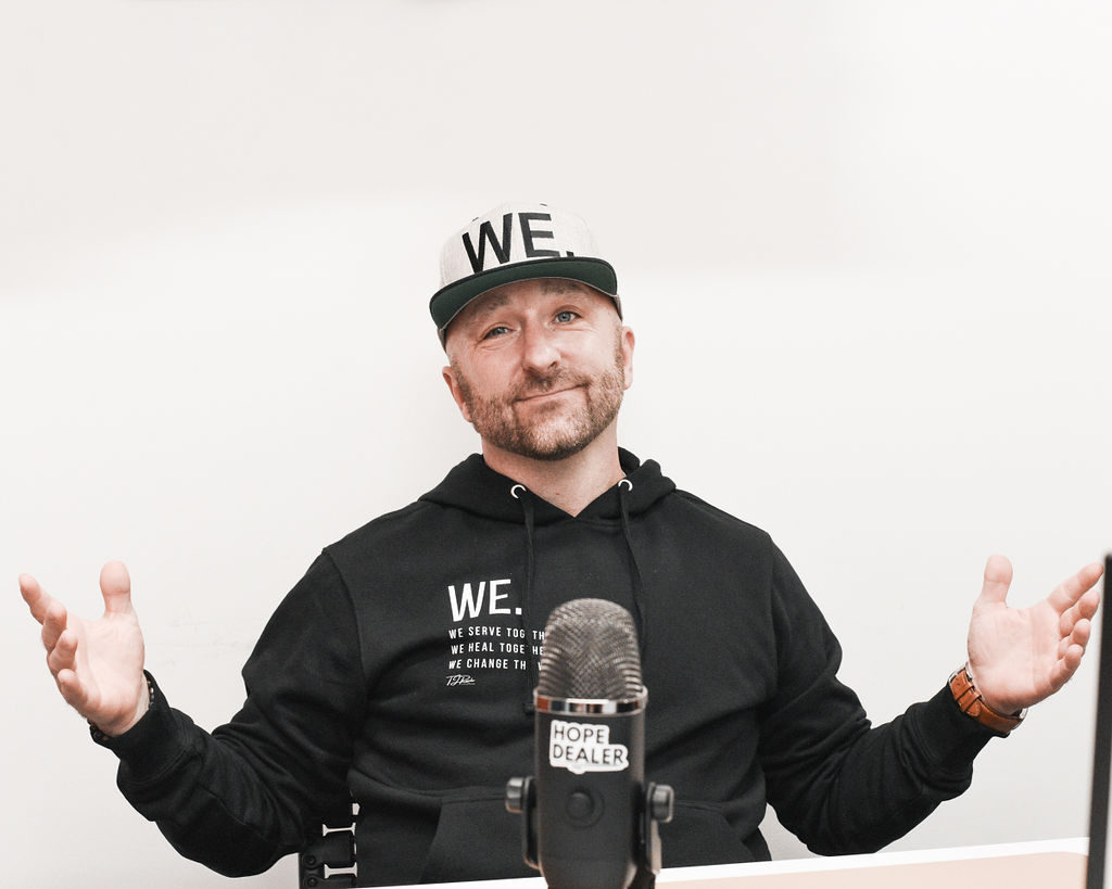 Man with beard wearing a black hoodie and cap with 'WE' on it, gesturing with hands, sitting in front of a microphone.